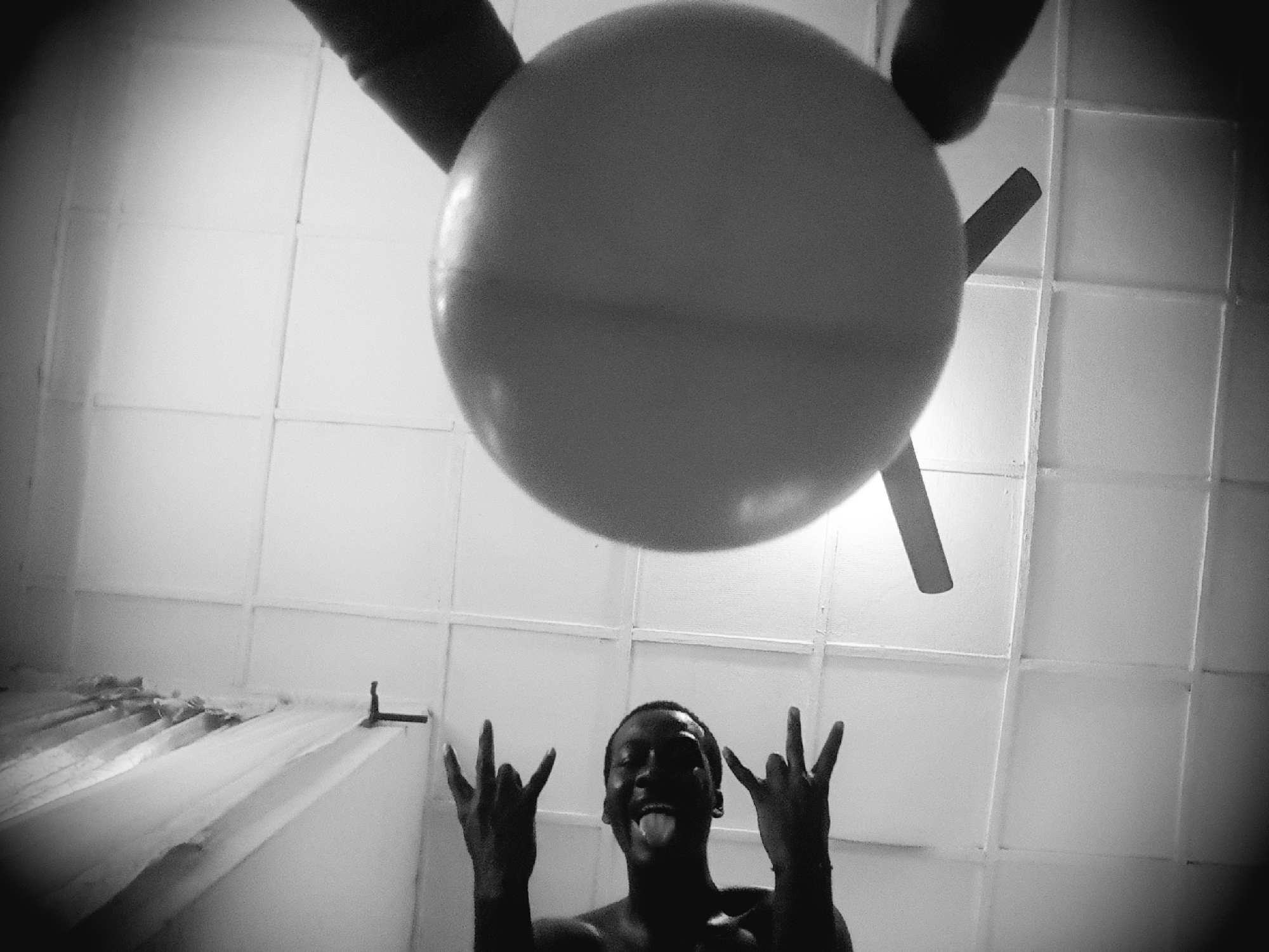 In an indoor environment, likely a bathroom, a low-angle shot looks up towards a tiled ceiling. A person, appearing to be male with dark skin, is positioned on the right side of the frame, looking upwards and extending both hands. His facial expression is obscured by the upward angle and shadows, but his mouth is open. His hands are raised, with fingers splayed in a gesture that could be interpreted as playful or threatening. On the left, a round, light-colored object, possibly a balloon, is partially visible, held by an unseen hand or resting against something. A ceiling fan with two blades is also visible, one blade extending towards the balloon. The overall lighting is dim, with some light emanating from the ceiling. The tiled surface of the ceiling is clearly defined by grout lines. A portion of what appears to be a shower curtain is visible along the bottom right edge of the frame.