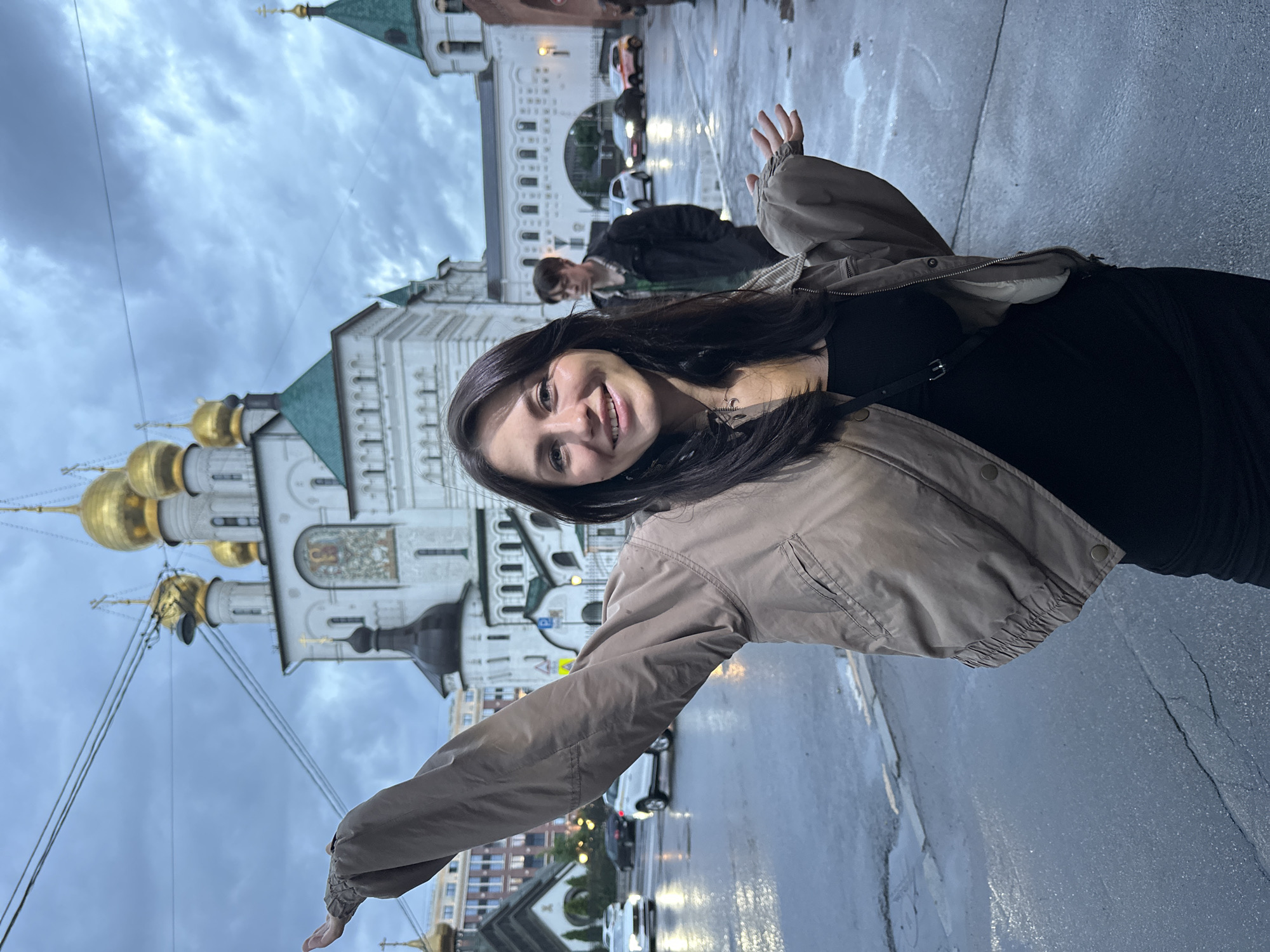 The image shows a young woman standing in front of a building with golden domes, likely a Russian Orthodox church, in Saint Petersburg, Russia. The weather appears to be overcast and the ground is wet, indicating recent rain. The woman is smiling and has her arms outstretched, as if in greeting or celebration. She is wearing a light-colored jacket over a black top and a black skirt. In the background, a young man is visible, along with several cars on a wet street. The architecture suggests a historical setting, common in Saint Petersburg. The lighting is dim due to the cloudy conditions.