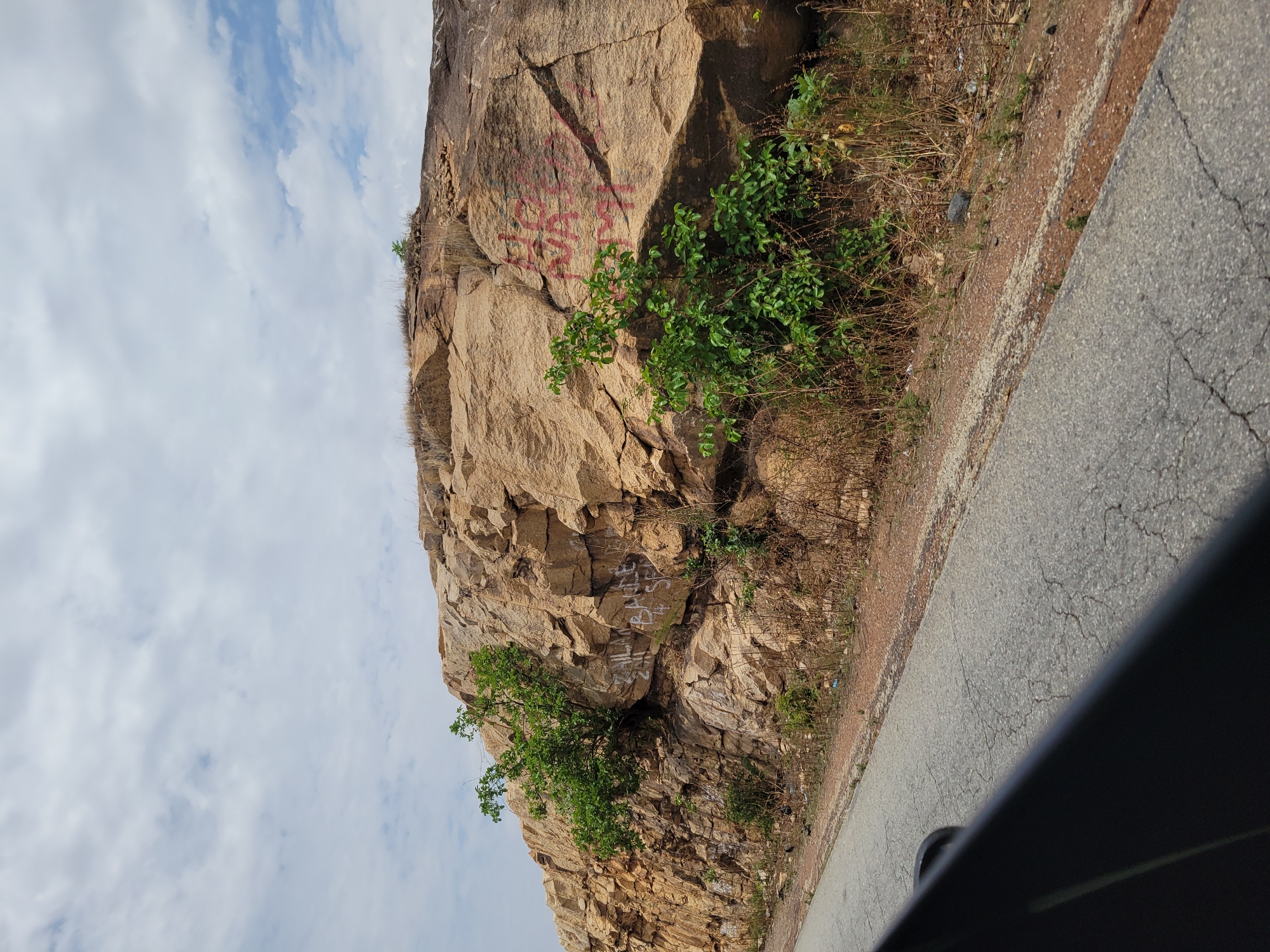 A photograph taken in Rimin Ziem, Nigeria, shows a large, tan-colored rock formation adjacent to a cracked asphalt road. The rock face is marked with reddish-pink graffiti and interspersed with green vegetation and dry grasses. A partially obscured section of what appears to be a dark car interior is visible in the lower right corner of the frame. The sky above is visible and is filled with scattered clouds.