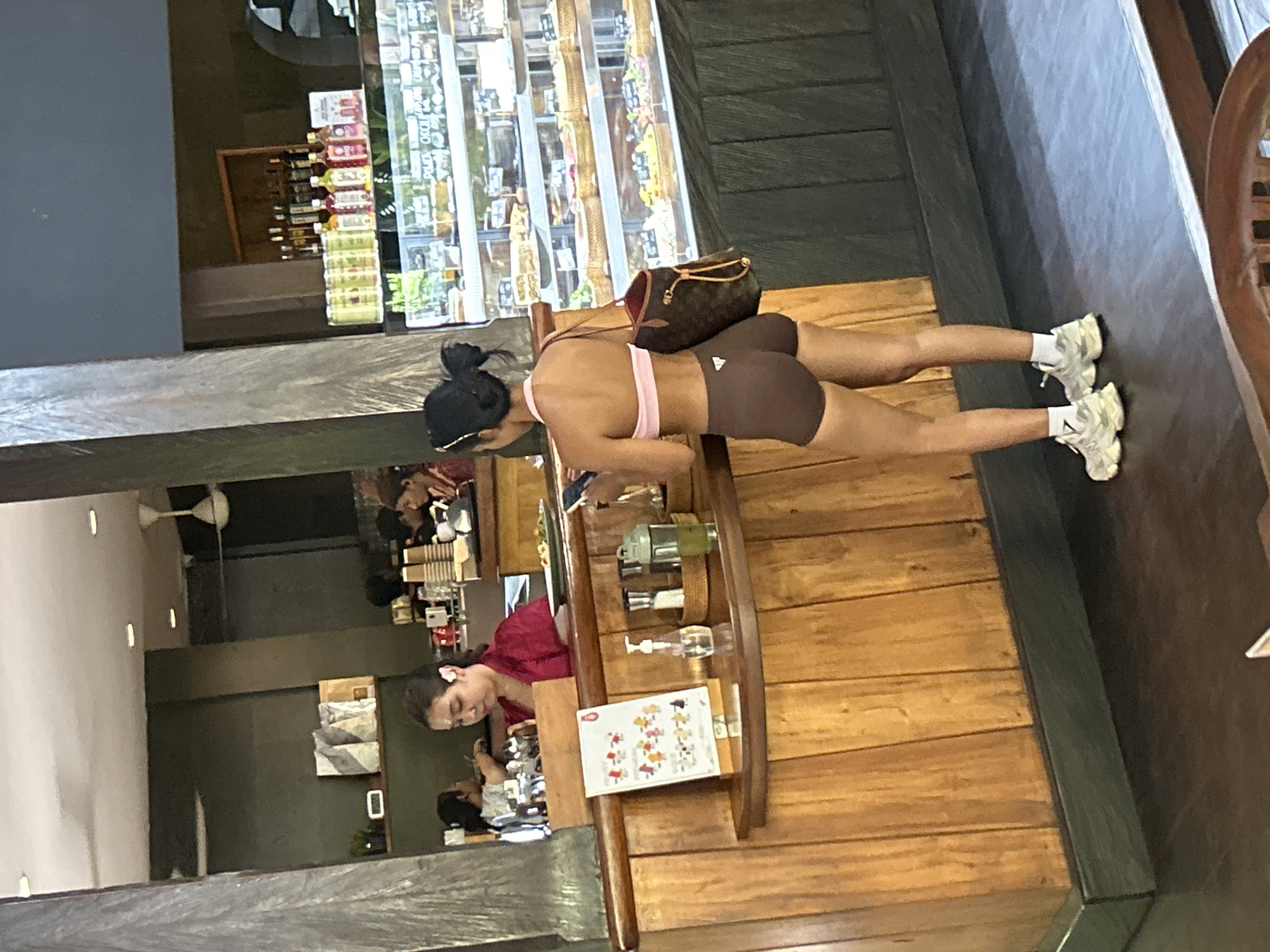 In North Kuta, Indonesia, a woman stands in a cafe, looking at her phone. She has dark hair pulled back in a bun and wears a pink sports bra and brown shorts. A brown designer bag is slung over her shoulder. In the background, two women in maroon shirts are working behind a counter, and behind them, shelves are stocked with various drinks and products. A refrigerated display case filled with pastries and desserts is visible to the right. The cafe features warm wood accents and dark flooring.