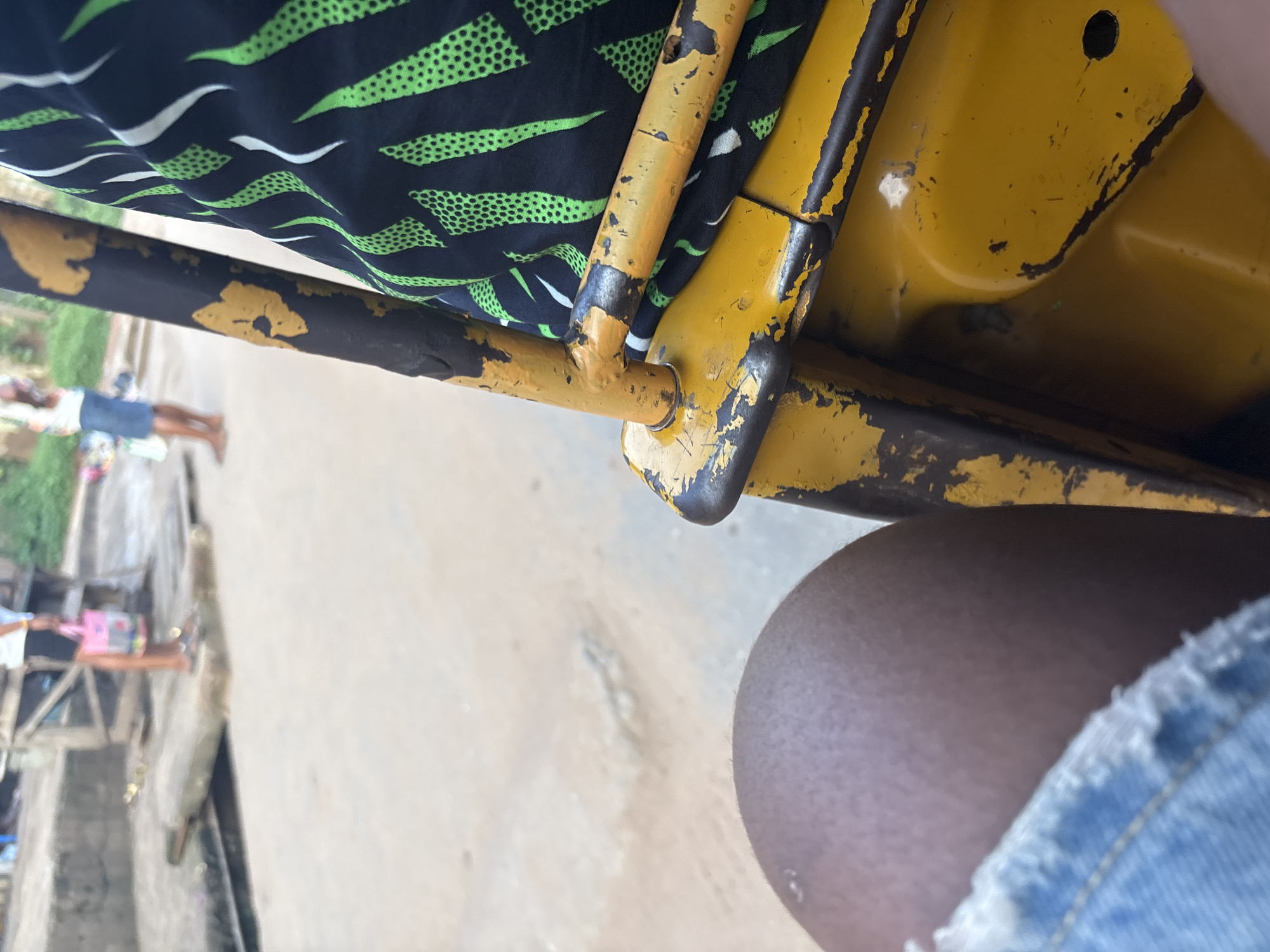 The image shows a close-up perspective of a person sitting in what appears to be a motorized tricycle, commonly known as an "okada" or "keke napep" in Nigeria. The seat is occupied by a person's legs, with part of their dark skin visible and a portion of their blue denim shorts with frayed edges in the lower left. The tricycle's yellow metal frame, showing signs of wear and peeling paint, is prominent in the foreground and to the right. A black fabric with a green abstract pattern is draped over the frame, obscuring part of the interior.
In the background, the scene is blurred but suggests an outdoor urban environment. Several people are visible, some standing and others appearing to be in motion. One person in the background on the left is holding a pink bag. The ground appears to be paved or dirt. The overall impression is that of being a passenger in a mode of transport in a busy, possibly market or street setting in Onitsha, Nigeria.