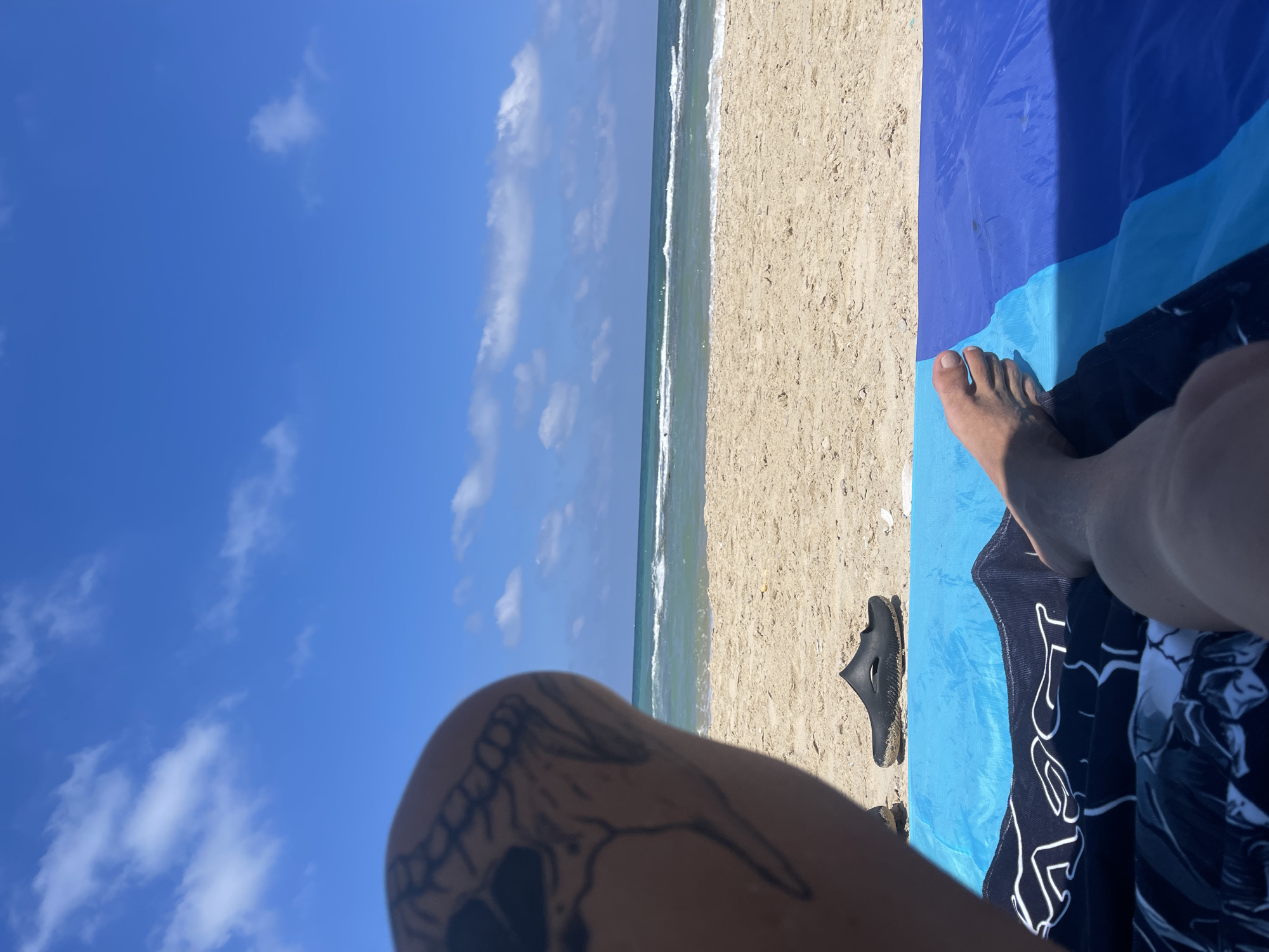 A first-person perspective shot captures a person relaxing on a beach in Ga'ash, Israel. The image focuses on the person's leg with a skull tattoo on their thigh, and their bare foot resting on a blue beach towel. A black sandal is placed on the sand nearby, with the ocean and sky visible in the background. The sea is calm with gentle waves, and the sky is bright blue with scattered white clouds, indicating a sunny day. The sand is light brown and appears dry.