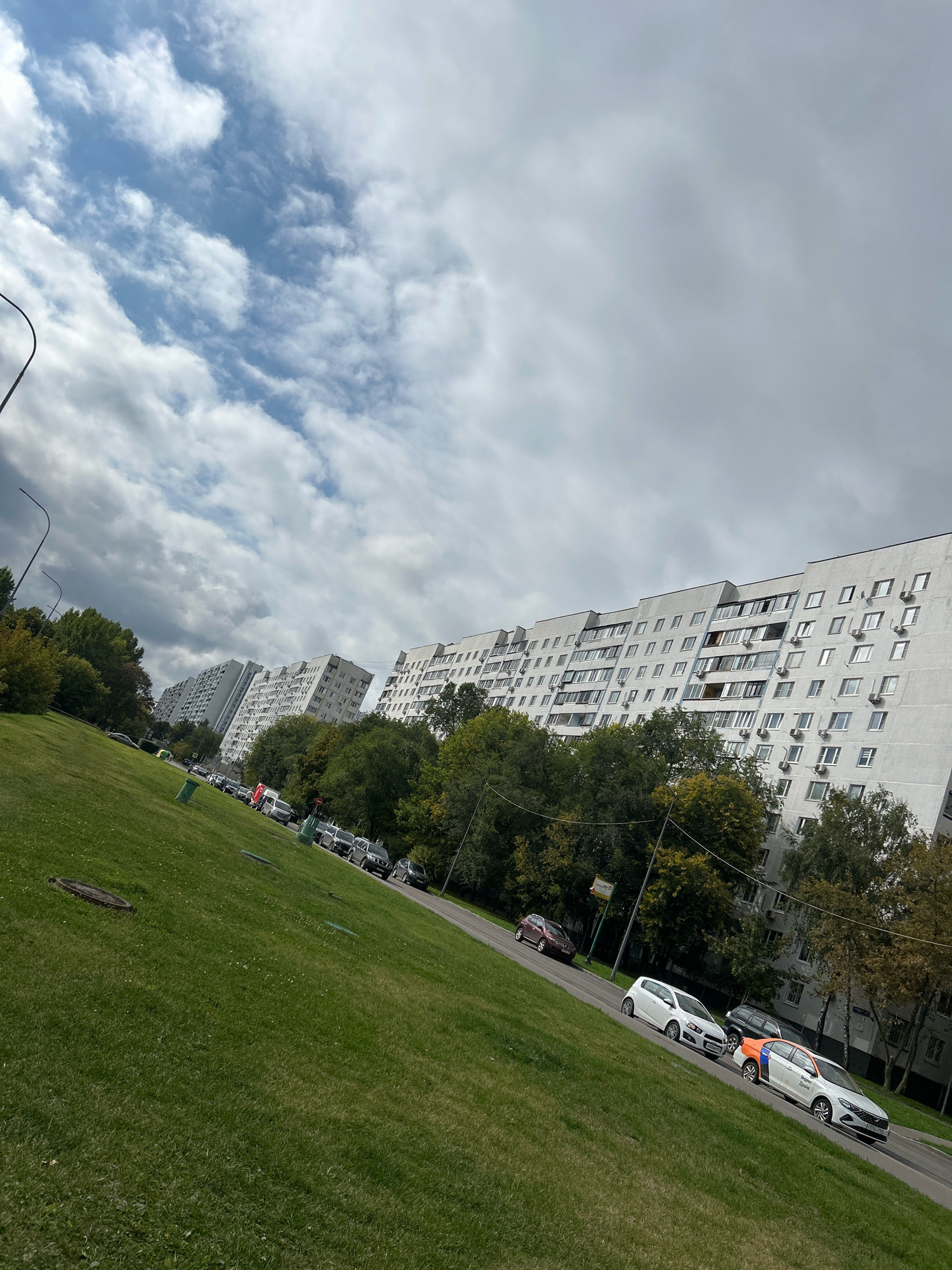The image captures a wide shot of a residential area in Moscow, Russia, likely on a cloudy day. In the foreground, a well-maintained, sloping green lawn with scattered leaves leads towards a road. Several cars are parked along the road, with some driving. Further back, a line of multi-story apartment buildings with numerous windows and air conditioning units stretches across the frame, characteristic of Soviet-era architecture. Dense green trees flank the apartment buildings, some showing hints of autumn foliage. The sky above is filled with dramatic, textured clouds. A street lamp is visible on the left side of the frame. The overall impression is one of a typical urban residential neighborhood.