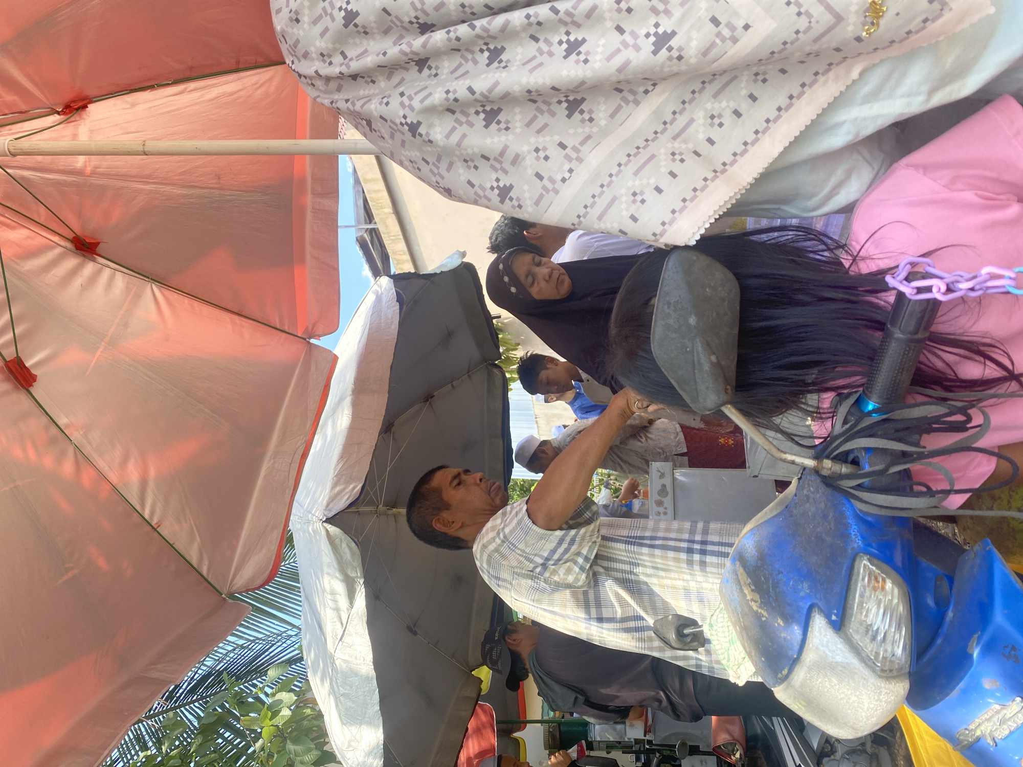In Banjarbaru, Indonesia, a street vendor, a man in a checkered shirt, is seen under a large pink umbrella, attending to customers. He is partially obscured by the umbrella and a blue scooter. The scooter's handlebars have black and grey straps hanging from them. To the right, a woman wearing a black hijab and a patterned headscarf is partially visible. Behind the vendor, other individuals are present, including a person in a dark hooded jacket and another wearing a white cap. The environment appears to be an outdoor market or a roadside stall, with other umbrellas and structures visible in the background, suggesting a bustling commercial area. The sky is clear and blue.