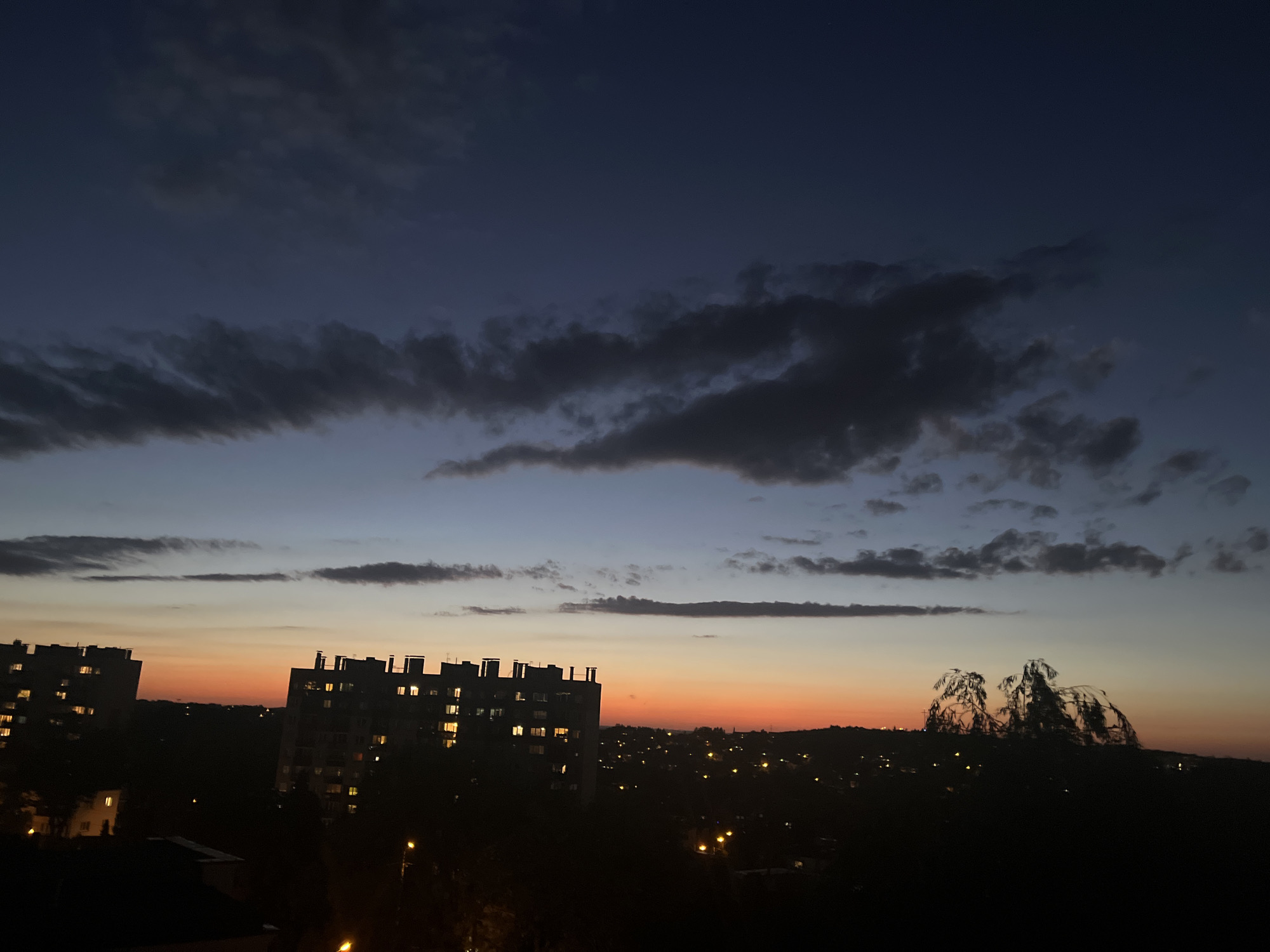 A wide, horizontal, eye-level outdoor shot captures a dusky cityscape with apartments and silhouetted trees against a gradient sky. The sky transitions from a deep, dark blue at the top to lighter blues and purples, then to a band of orange and yellow near the horizon, before fading into the dark silhouettes of the city below. Several clouds are visible in the sky, with darker, more defined clouds in the upper portion and lighter, wispy ones closer to the horizon.
In the foreground and mid-ground, dark silhouettes of buildings and trees dominate. Two large apartment buildings are prominent, with many windows illuminated by warm, yellow light, suggesting residents are home. The apartment buildings appear to be of similar height and architecture, suggesting a residential development. To the right, silhouetted trees with intricate branches are visible against the horizon, adding a natural element to the urban scene. The rest of the cityscape is a dark, undulating silhouette punctuated by scattered points of light, indicating streetlights or other urban illumination. The overall impression is one of a quiet evening in a residential area of the city.