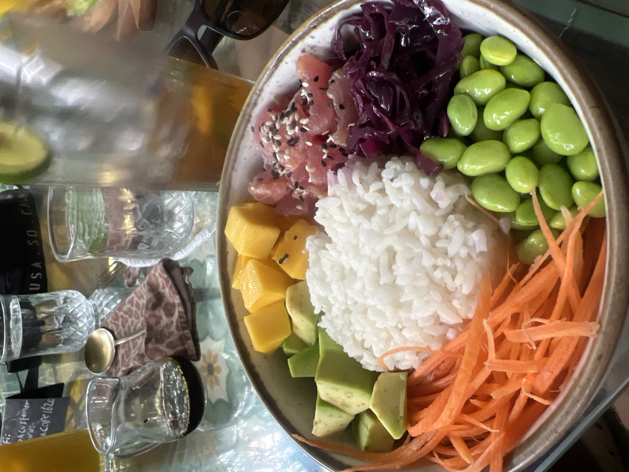 A close-up, overhead shot focuses on a poke bowl brimming with colorful ingredients on a reflective glass table. The bowl contains white rice, diced raw tuna seasoned with black and white sesame seeds, shredded red cabbage, edamame, julienned carrots, diced avocado, and diced mango.
In the background, other elements of a dining setting are visible. To the left, a small glass pitcher holds a dark liquid, possibly soy sauce or dressing, next to a patterned napkin with a golden spoon. Behind the pitcher, a glass of water sits on the table. To the right, another glass filled with a beverage containing ice, citrus slices, and possibly mint is partially obscured by the poke bowl. A pair of dark sunglasses rests on the table near the beverage. The table surface reflects the objects placed on it.
The location is identified as Abang, Indonesia, suggesting a restaurant or dining establishment within that region.