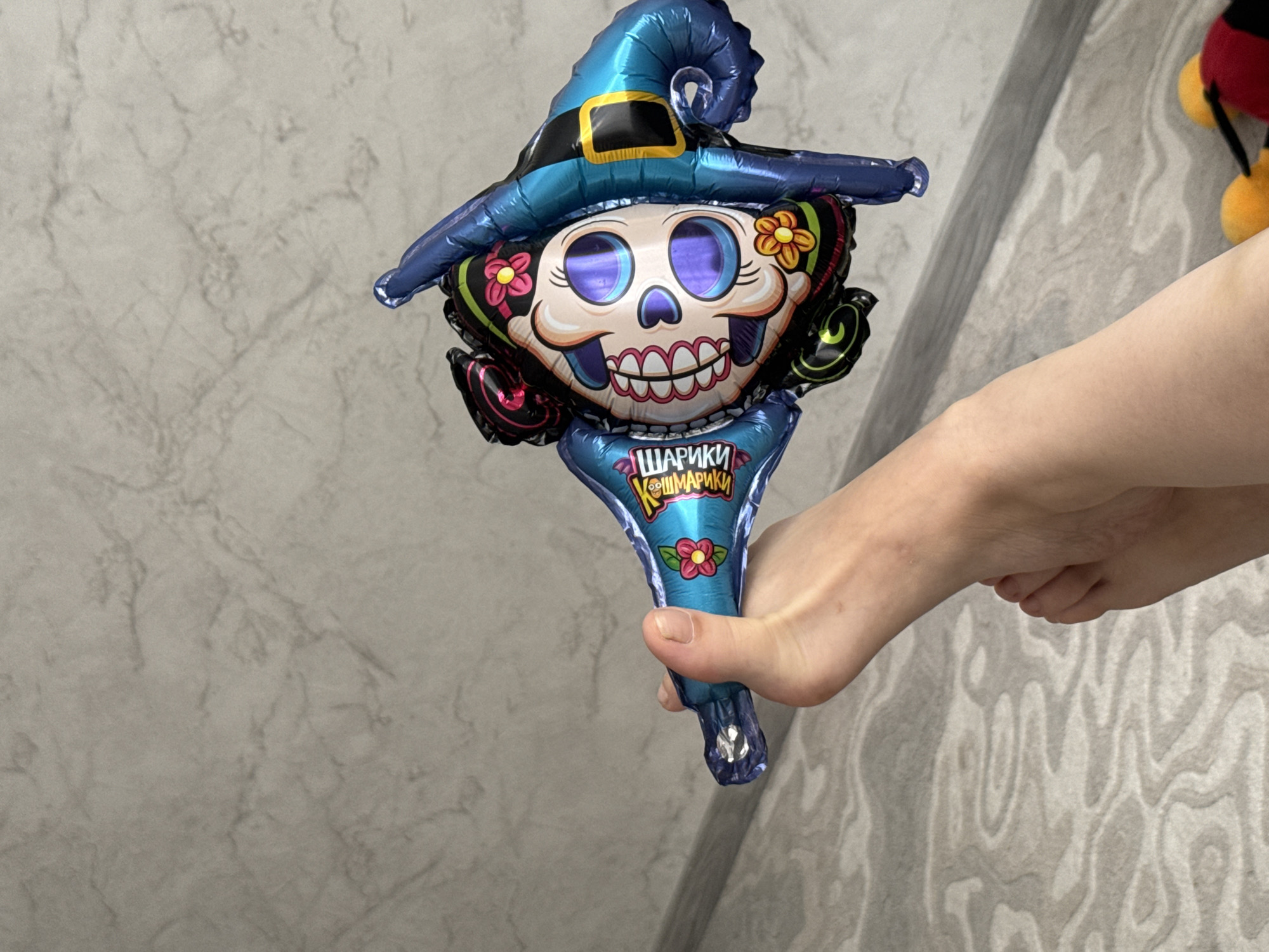 A person's hand is holding a balloon shaped like a skull with a witch's hat. The balloon is primarily blue and black with colorful decorations on the skull, including flowers and teeth. The person's foot is visible in the lower part of the image. The background consists of a textured wall and a patterned rug. The scene appears to be indoors. The provided location of Kostroma, Russia, is not discernible from the visual elements of the image.