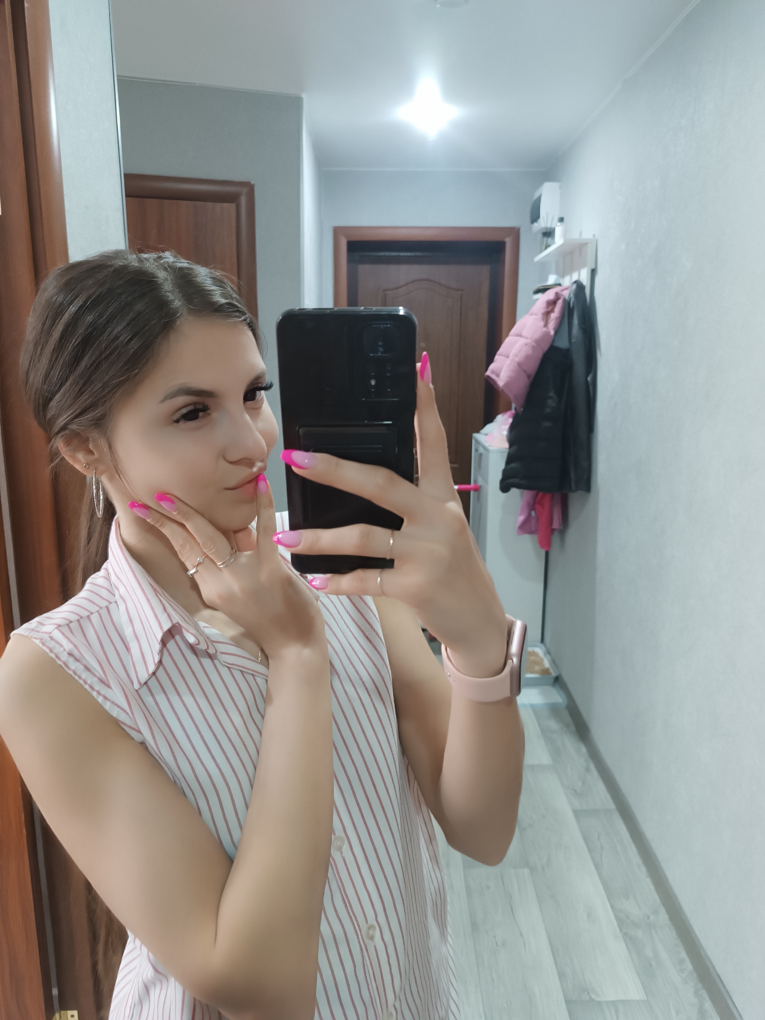 A young woman with brown hair pulled back and bright pink acrylic nails takes a mirror selfie in a hallway. She is wearing a white and pink striped sleeveless button-down shirt. On her left wrist, she wears a pink smartwatch. Her right hand is raised to her face, with her fingers touching her cheek and lips. In the background, a mirror reflects a hallway with a wooden door on the left, a white shelf with items on the right wall, and several jackets hanging on the wall. The floor is light-colored wood laminate.