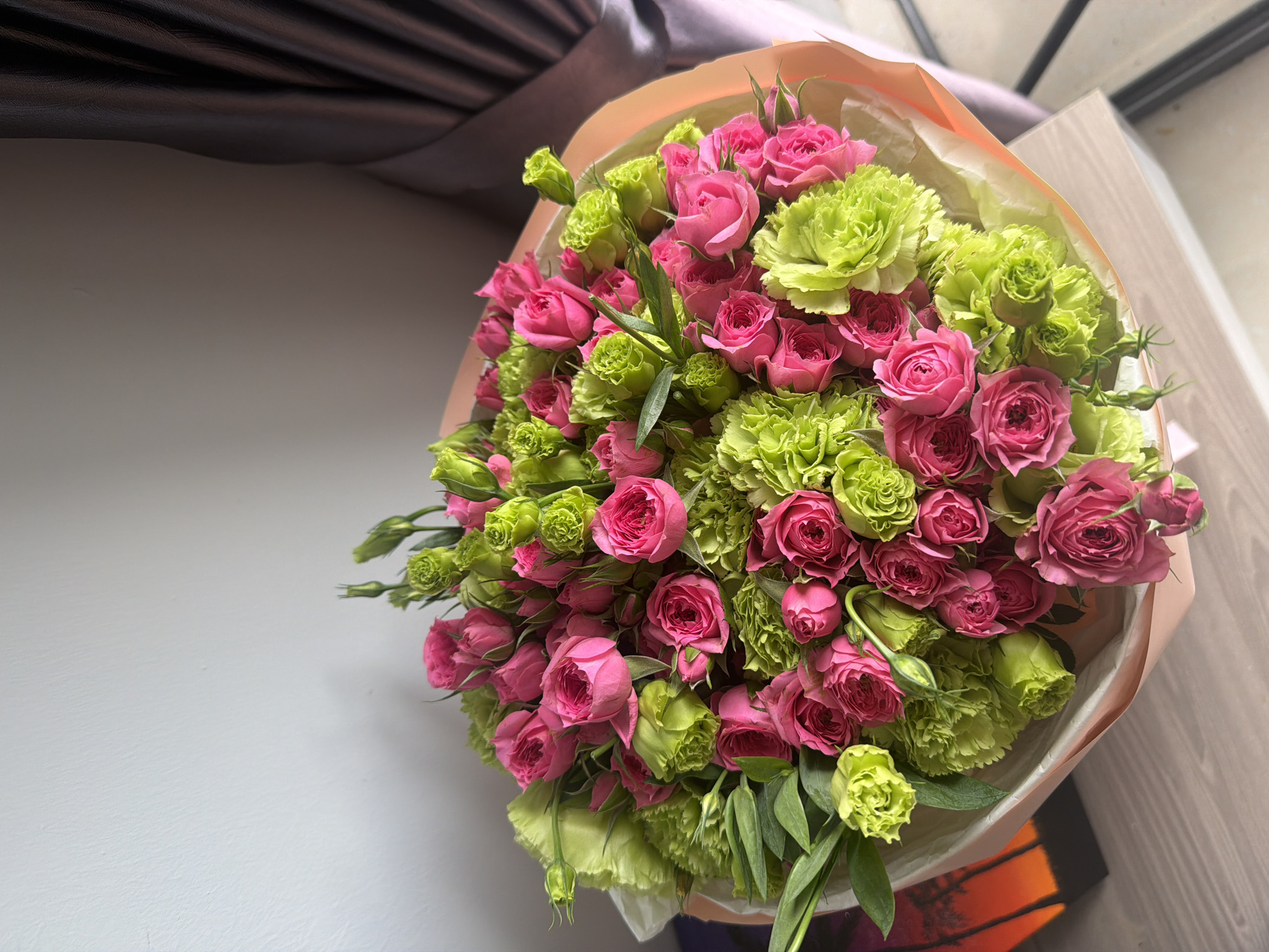 The image shows a bouquet of pink roses and green carnations arranged in a light pink paper wrapping. The bouquet is placed on a wooden surface, likely a table, against a plain white wall on the left and a dark gray curtain on the right. There are no people visible in the image, and no specific actions or interactions are depicted. The lighting suggests an indoor environment, possibly a room. The location of Nha Trang, Vietnam, is not identifiable from the visual elements present in the image.