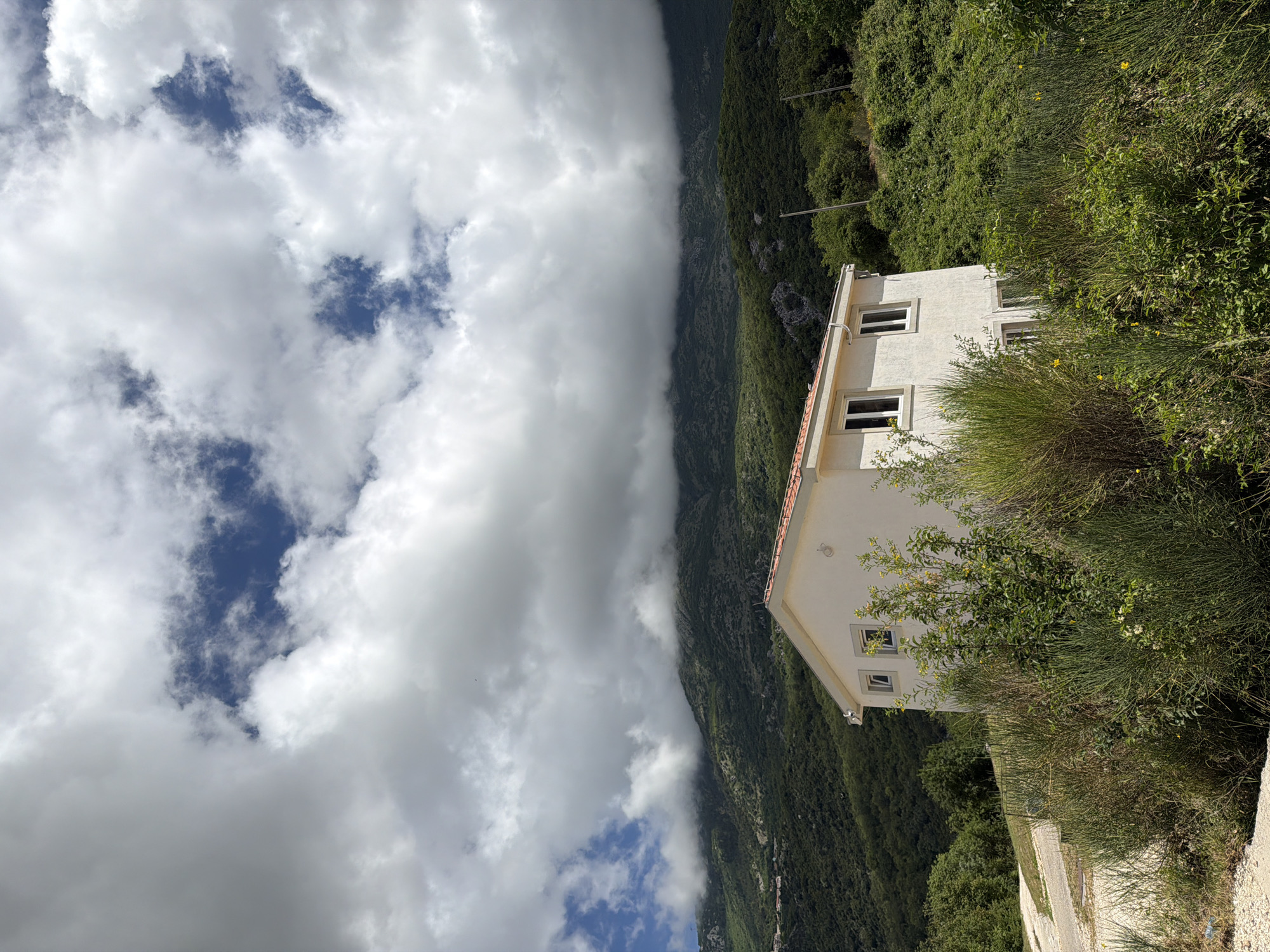 The image depicts a serene mountainous landscape in Bratešići, Montenegro, under a partly cloudy sky. In the foreground, a modern two-story white house with a reddish-brown tiled roof is partially obscured by dense, wild green vegetation, including tall bushes and trees. Several windows are visible on the house's facade. A light-colored dirt or gravel path runs along the bottom edge of the frame, leading towards the house.
Behind the house and extending across the midground, verdant mountains rise steeply, covered in a thick canopy of trees and foliage. A dramatic band of low-hanging, dark grey clouds stretches horizontally across the mountain range, partially obscuring the peaks and creating a sense of depth and atmosphere. Above these lower clouds, the sky features a mix of bright white and softer grey clouds, with distinct patches of clear blue sky visible.
The scene captures a peaceful, natural environment, with no discernible human activity or events taking place. The lighting suggests it is daytime, possibly morning or early afternoon, with ample natural light illuminating the landscape. The weather is dynamic, showcasing both clear skies and significant cloud cover. There is no visible text in the image.
