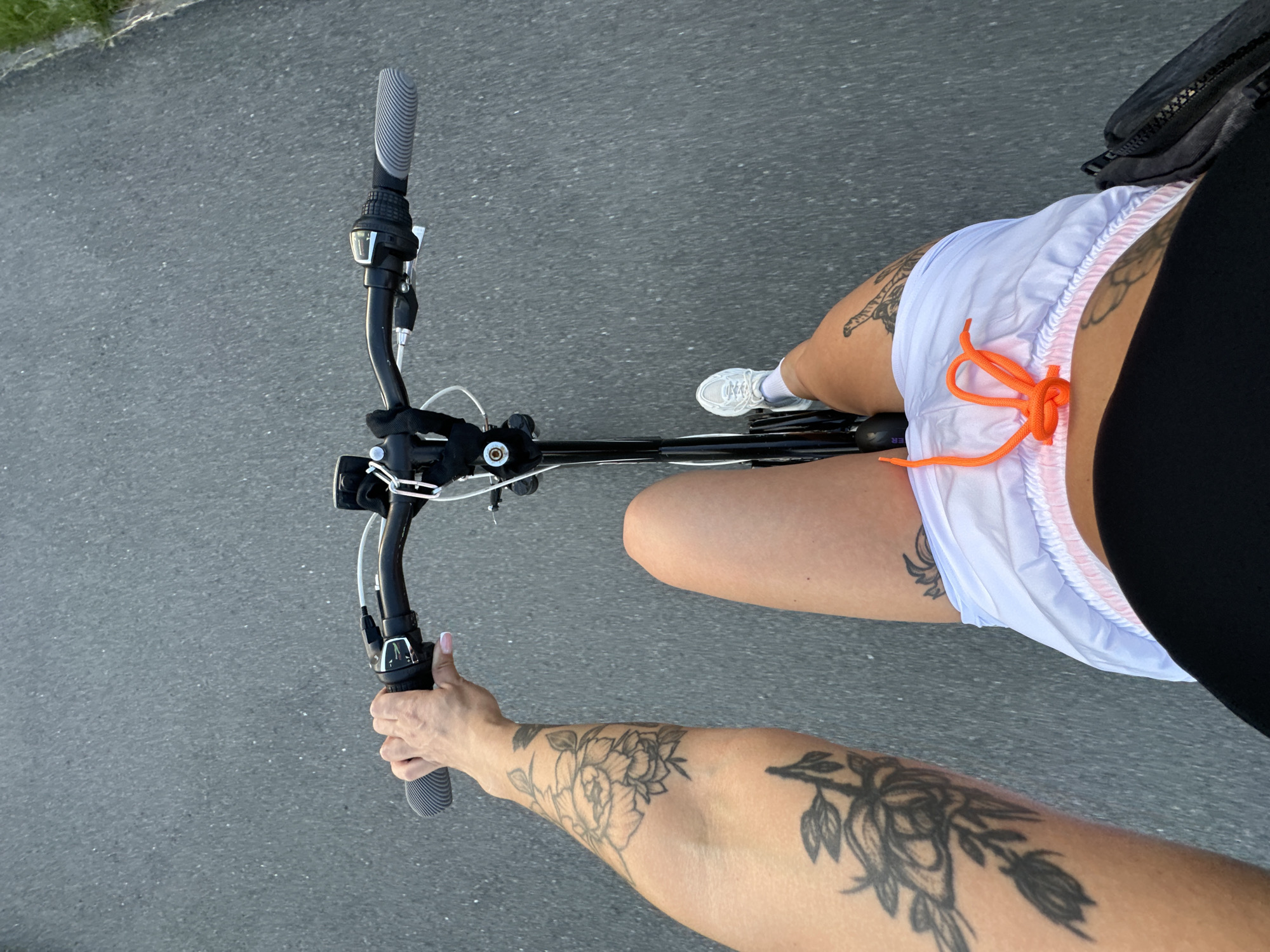 This is a first-person perspective, point-of-view shot of a person riding a bicycle on an asphalt path. The rider's left arm, adorned with floral tattoos, is visible holding the handlebars. Their legs are also visible, wearing white athletic shorts with an orange drawstring and white sneakers. The background consists of the asphalt path and a grassy area on the right edge. The scene appears to be outdoors, possibly in a park or recreational area. No specific landmarks identifying Yuzhnouralsk, Russia, are present.
