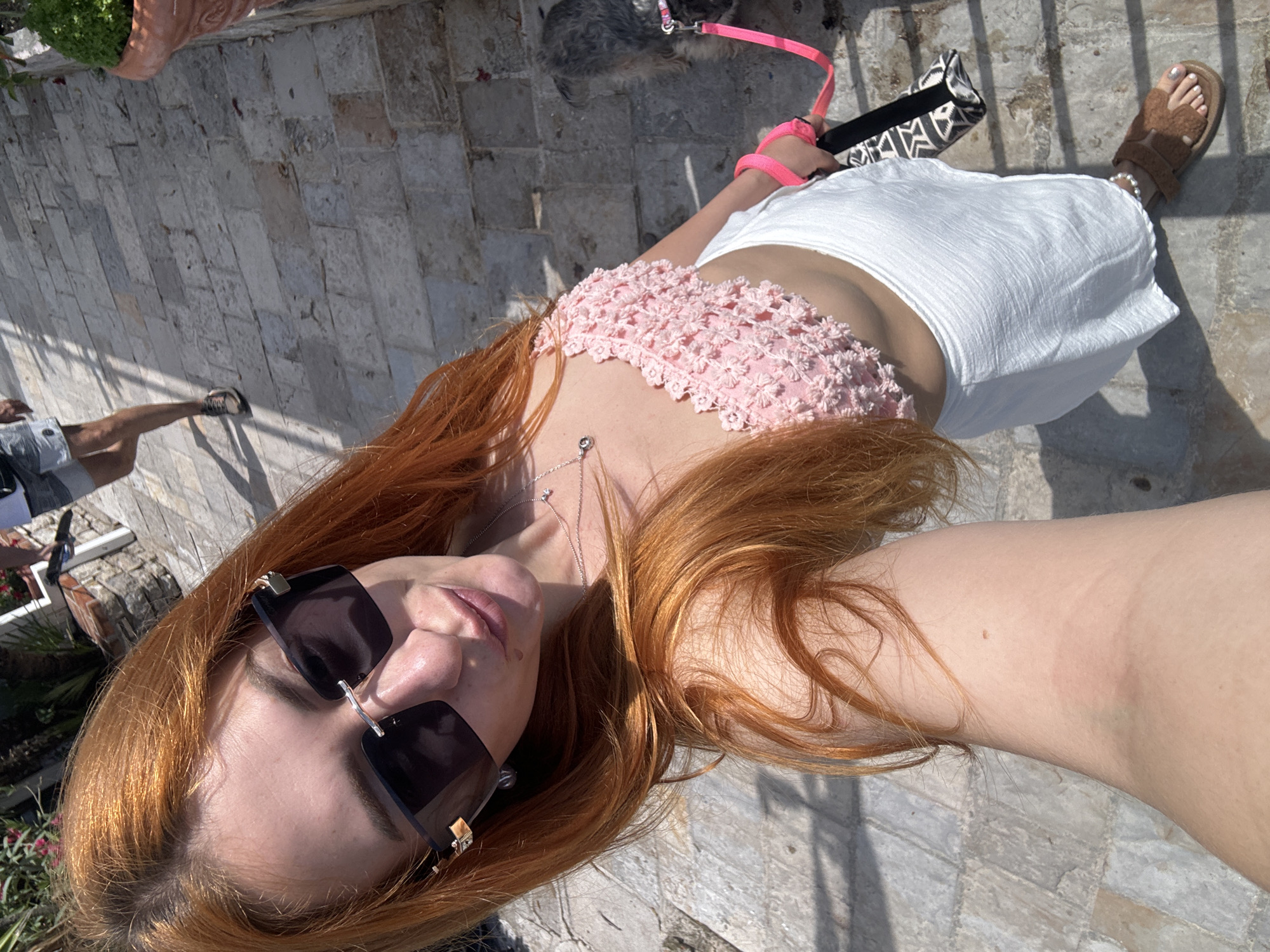 A high-angle, slightly rotated selfie captures a woman outdoors on a bright, sunny day, likely in Budva, Montenegro. The woman, with long, coppery-red hair, is wearing dark, rectangular sunglasses, a light pink crocheted crop top with floral-like details, and a white, textured skirt. She has a silver necklace with a small pendant. In her right hand, she holds a bright pink dog leash, which connects to a small, fluffy gray and black dog visible nearby. She also holds a black and white geometric-patterned clutch bag. One of her feet, adorned with white painted toenails, is visible in a brown, fluffy sandal with a white beaded ankle bracelet.
The setting appears to be an outdoor paved area, possibly a walkway or patio, made of light-colored, rectangular flagstones. Strong, distinct shadows are cast across the ground, indicating bright sunshine and clear weather, likely late morning or early afternoon. In the background, the lower legs of another person wearing dark shorts and sneakers are visible, suggesting companionship. Hints of greenery from potted plants or landscaping are also present. No visible text is present in the image. The scene conveys a relaxed, casual atmosphere during an outdoor activity.
