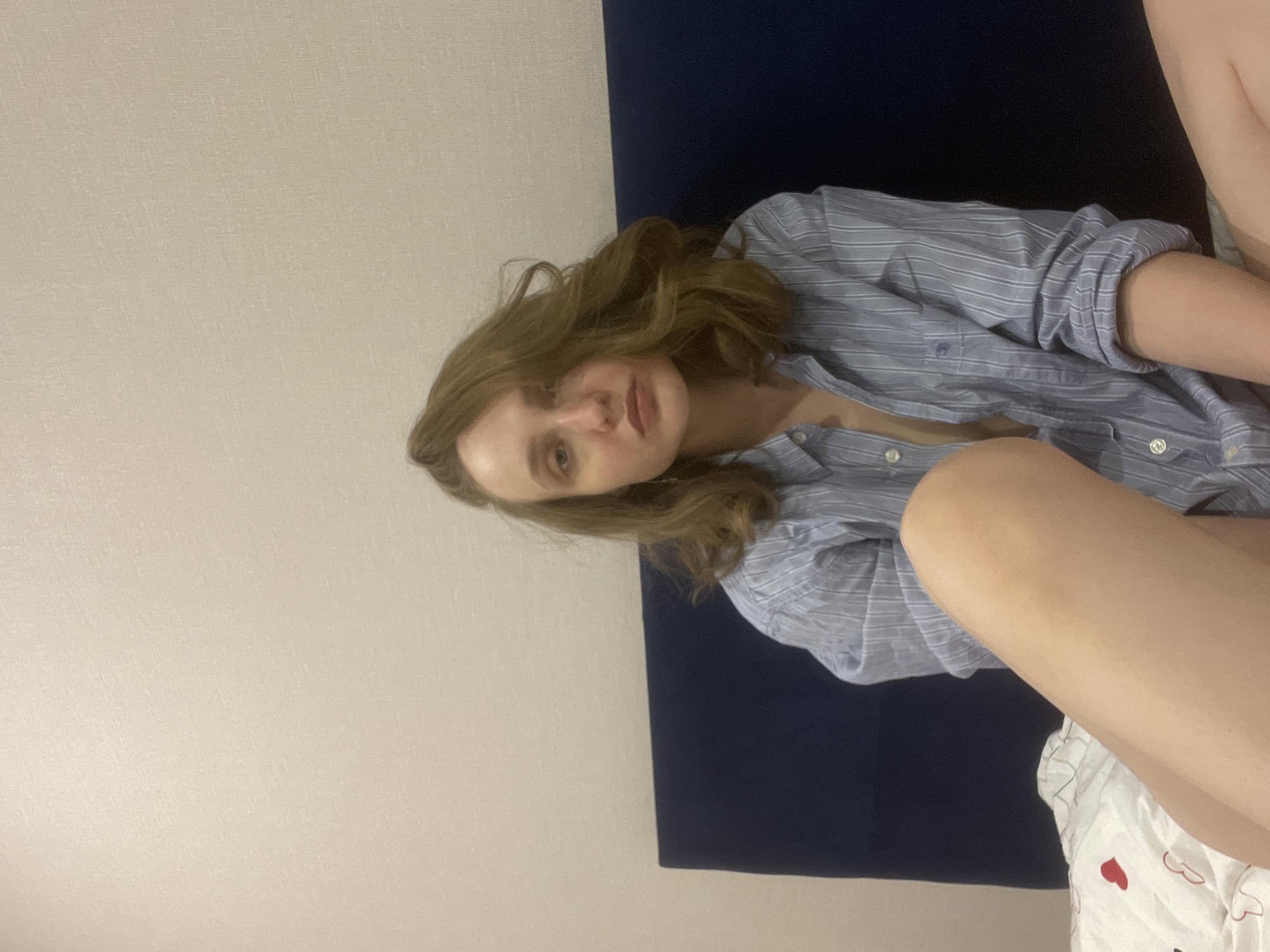 A young woman with wavy, light brown hair is pictured in a casual indoor setting. She is wearing a loose-fitting, light blue and white striped button-down shirt with rolled-up sleeves, and several top buttons are undone. Her expression is neutral as she looks directly at the camera. She appears to be reclining or sitting in a relaxed posture, with her bare arm and leg visible. Behind her is a dark blue surface, possibly a bed headboard or couch backrest, and a plain, light beige wall. A white blanket or duvet adorned with small red heart patterns covers part of her lower body in the bottom right of the frame. The lighting is soft. No text is visible. The image was taken in Podshivalovo, Russia.