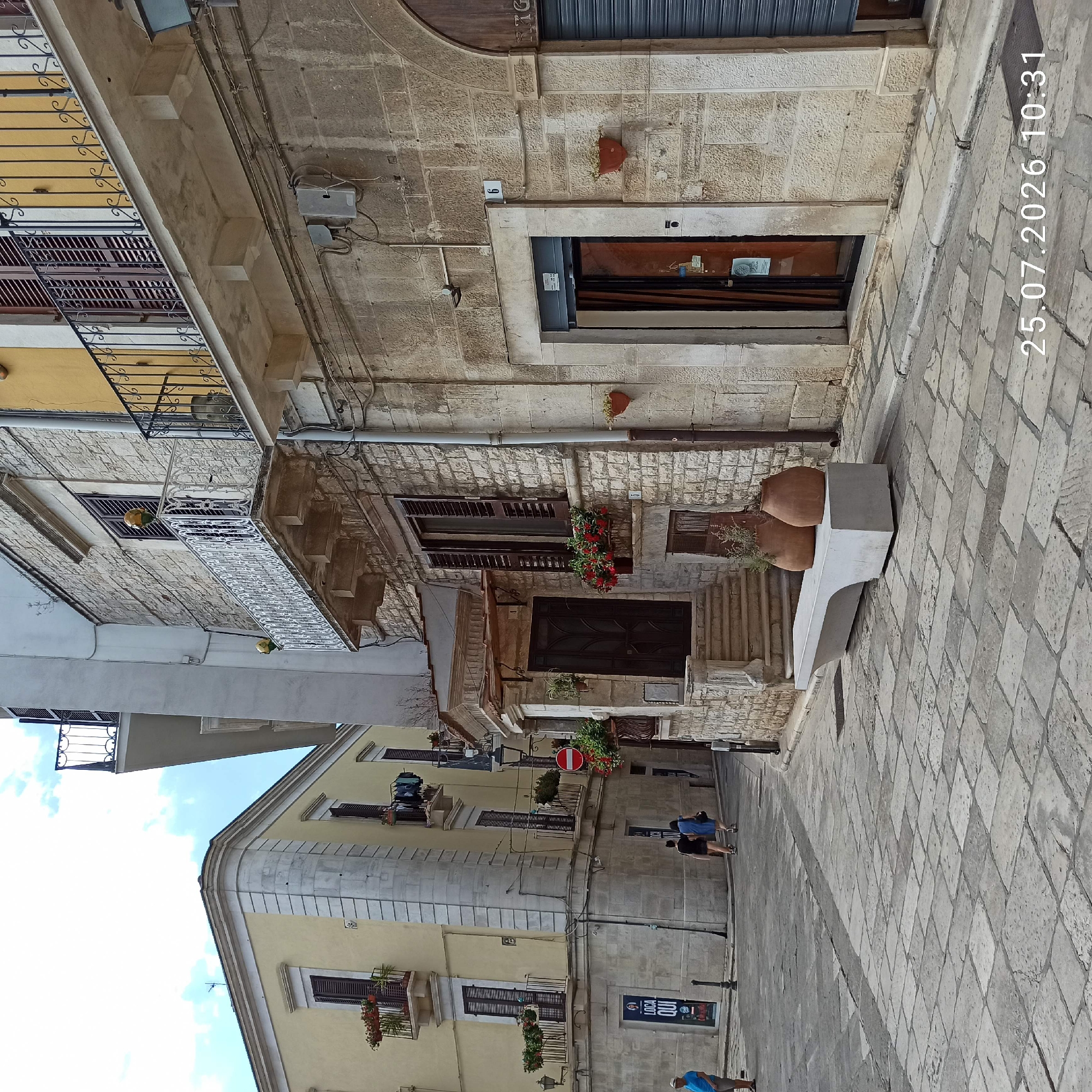 This is a street scene in Ruvo di Puglia, Italy. The image features cobblestone paving and stone buildings with balconies and shutters. A concrete bench with two large terracotta pots sits in the foreground. To the left, a pale yellow building has balconies with flowers. To the right, a stone building has a doorway with a dark wooden door and a metal roll-up shutter. The date stamp "25.07.2026 10:31" is visible in the lower right corner. Two women are walking away from the camera down the street on the left side of the frame, and a man in a blue shirt and a straw hat stands on the far left.