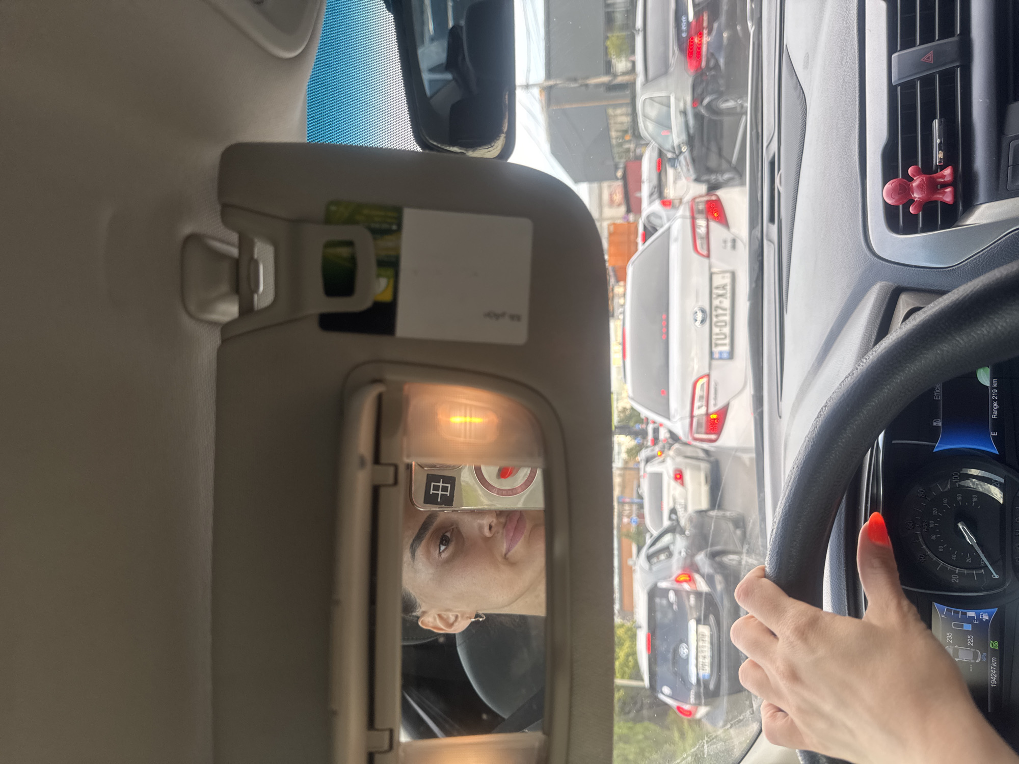 This media file captures a view from inside a car, likely in Tbilisi, Georgia, during the daytime. The scene depicts a traffic jam, with multiple vehicles visible through the windshield. The sun appears to be shining, suggesting fair weather.
The primary subjects are a person's hands on the steering wheel and a reflection of the person's face in the car's vanity mirror. The driver has bright orange nail polish. The vanity mirror is illuminated by a light and shows a person's face, with a glimpse of a character symbol (中) on a sticker or phone case.
Notable details include the car's dashboard with a speedometer displaying speed in both MPH and km/h, and an odometer reading of 194,247 km. A license plate with the text "TU-017-XA" is visible on one of the cars in traffic. An air freshener in the shape of a red figure is clipped to a car vent. The image also shows a sun visor on the left side of the frame. The overall impression is of someone waiting in traffic, possibly looking at themselves in the mirror.