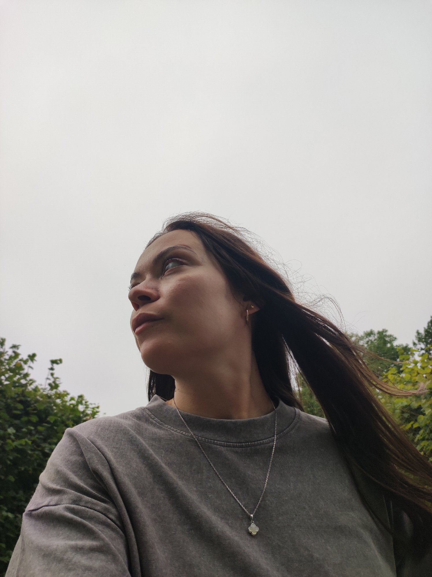 A low-angle, medium shot shows a young woman from the chest up, looking up and to the left. She has long, dark brown hair that is blowing to the right, obscuring part of her face and neck. She is wearing a gray crewneck sweatshirt and a silver necklace with a four-leaf clover pendant. Her left ear has a small gold hoop earring. She has light skin and appears to be outdoors, with blurred green foliage in the background and a pale, overcast sky above.