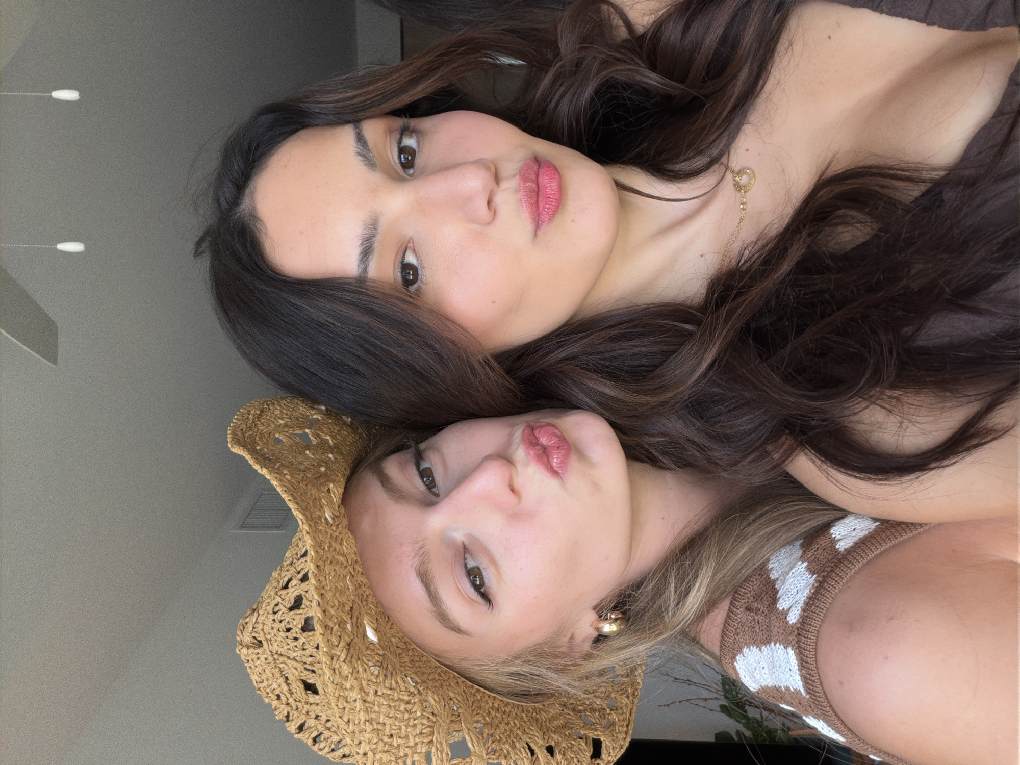 Two young women pose for a selfie. The woman on the left wears a straw cowboy hat and a brown and white patterned top. She has blonde hair and is puckering her lips. The woman on the right has dark, wavy hair and is also puckering her lips. She wears a dark-colored top and a gold necklace. The background appears to be an indoor setting with a ceiling fan visible. The image is cropped to focus on the faces and upper bodies of the two women.