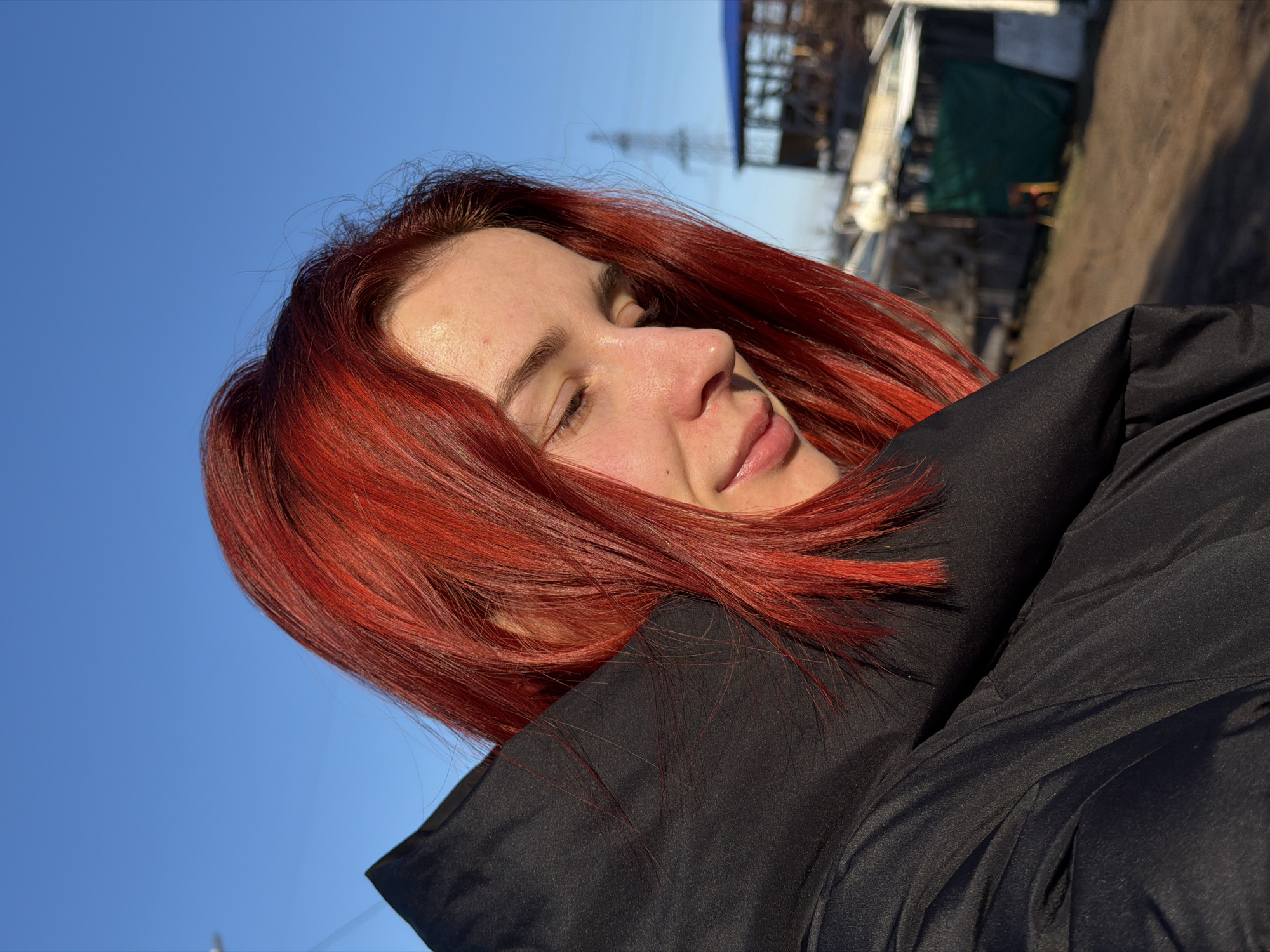 A medium, angled shot shows a young woman with dyed red hair, light skin, and closed eyes, appearing serene. She is wearing a dark, puffy jacket. In the blurred background, there are urban structures, utility poles, and possibly industrial buildings under a blue sky, which suggests the location is Chaikovsky, Russia, based on known city characteristics. The image is taken during daylight, with clear lighting conditions.