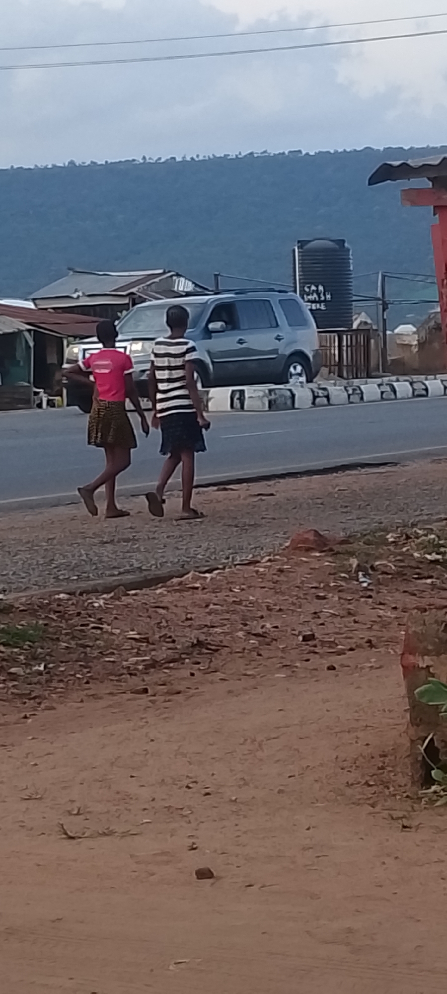 The image captures a street scene in Adankolo, Nigeria. Two children are walking alongside a paved road, one wearing a pink shirt and patterned skirt, the other a striped shirt and dark skirt. Both are wearing sandals. Behind them is a grey SUV parked near a curb painted with alternating black and white stripes. Further back, there are structures including buildings with corrugated roofing and a large black water tank labeled "Car Wash". In the background, a blue-tinged mountain range is visible under a cloudy sky. The foreground shows a dirt shoulder along the road with reddish-brown soil and scattered debris. A corner of a red post is also visible.