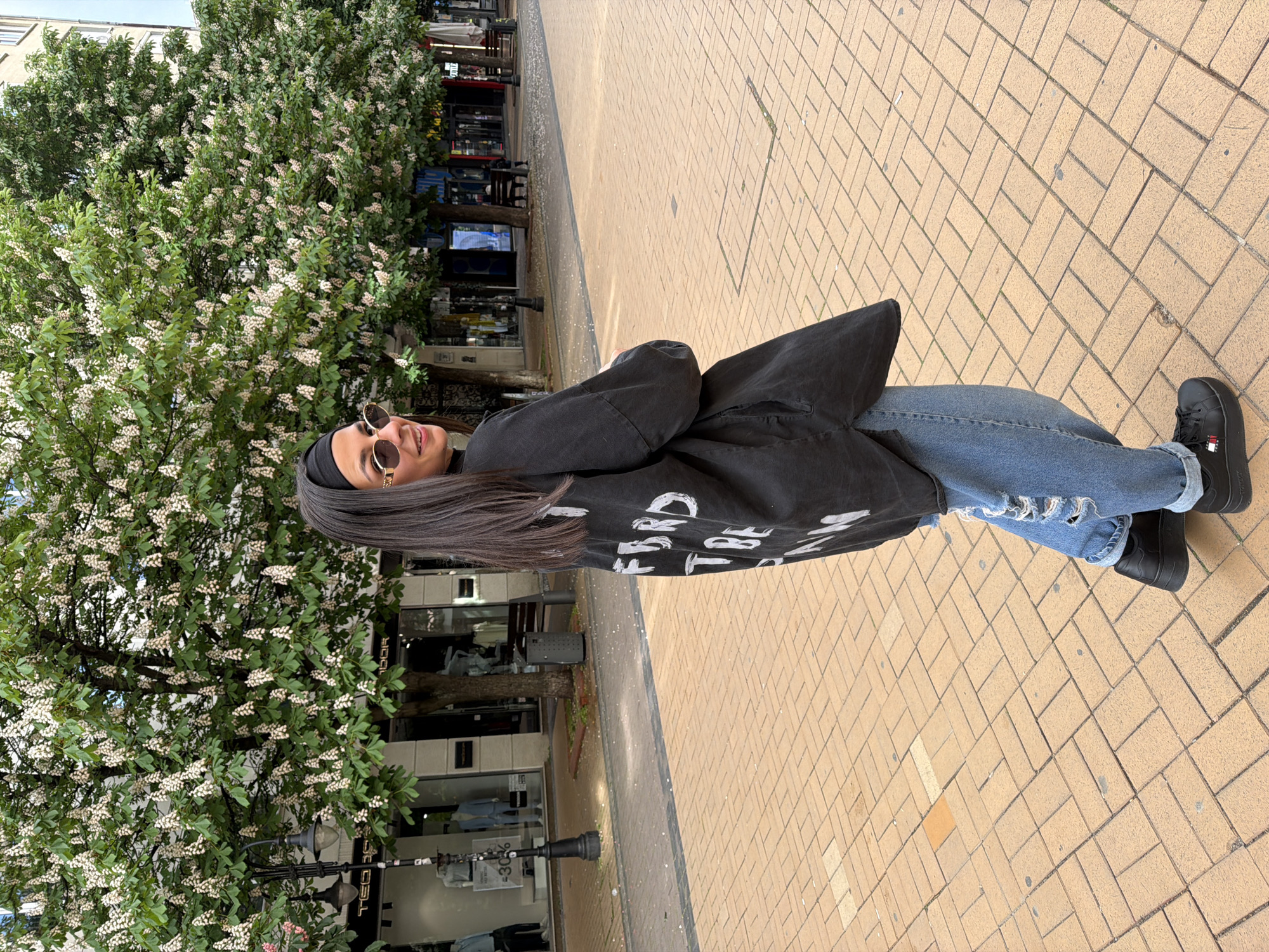 A woman stands on a brick-paved sidewalk in Sofia, Bulgaria. She wears a black head covering, sunglasses, a black denim jacket with white lettering, blue jeans, and black shoes. She is positioned in front of a tree filled with white flowers. Behind her is a street with shops and street furniture.