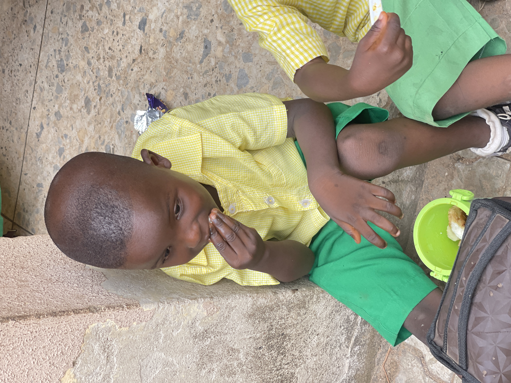 In Akwanga, Nigeria, a young child, likely a boy, is seated on a paved surface next to a textured, light-colored wall. He is wearing a yellow and white checkered collared shirt and green trousers. The child is bringing a piece of food to his mouth with his right hand. Next to him, another child, also in a similar yellow and white checkered shirt and green trousers, is partially visible. In front of the first child is a small, bright green plastic container holding a piece of bread. A foil wrapper, likely from candy, lies on the ground nearby. A dark-colored bag with a patterned texture rests beside the child's leg. The ground is composed of small, light-colored stones and concrete.