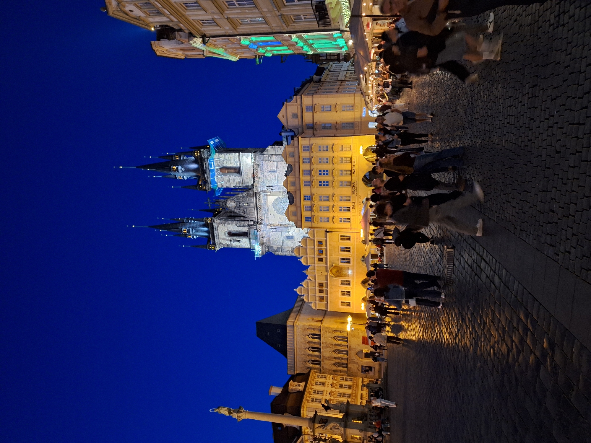 The image captures a bustling nighttime scene at the Old Town Square in Prague, Czechia. Dominating the background is the iconic Church of Our Lady before Týn, its Gothic spires illuminated against the deep blue twilight sky. To its left stands the Jan Hus Memorial and a column. Surrounding the square are historic buildings with warm yellow lighting emanating from windows and street lamps, reflecting on the cobblestone pavement.
In the foreground, numerous people are walking and gathered in the square. Many are casually dressed, suggesting they are tourists or locals enjoying the evening. Some are walking towards the camera, while others are walking away or conversing in groups. The cobblestone ground is wet in places, creating shimmering reflections of the lights. The atmosphere appears lively and social, with outdoor seating and umbrellas visible from cafes and restaurants lining the square.