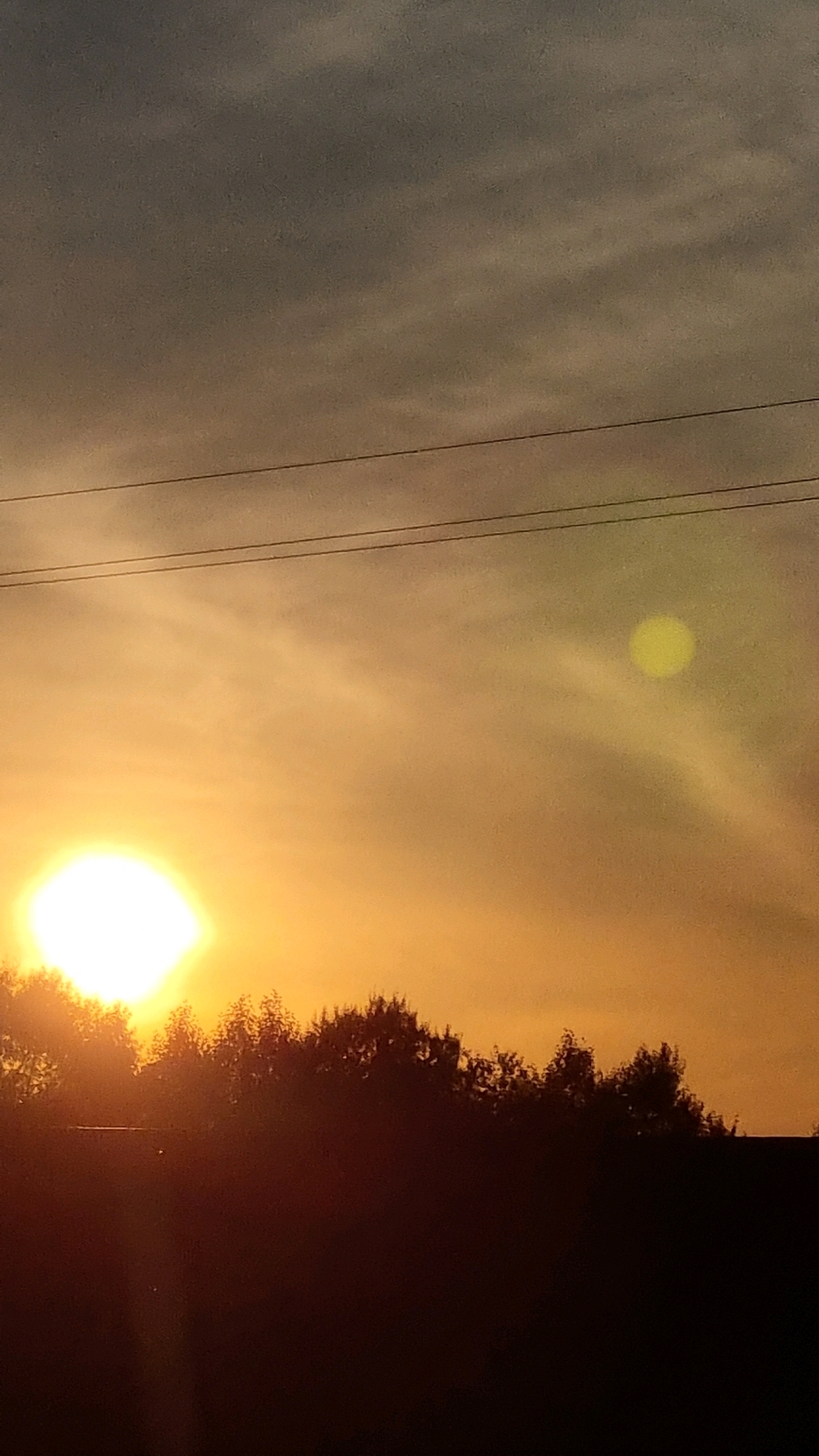 The image depicts a sunset over the city of Vysokoe, Russia. The sky is a gradient of warm orange and yellow hues, with darker gray clouds towards the top. Two parallel power lines stretch horizontally across the upper portion of the frame. The sun, a bright white disc, is partially obscured by a silhouetted treeline in the lower half of the image. A faint, circular yellow light artifact is visible in the upper right quadrant of the sky, likely a lens flare. The foreground is dominated by the dark silhouettes of trees and possibly a building structure.
