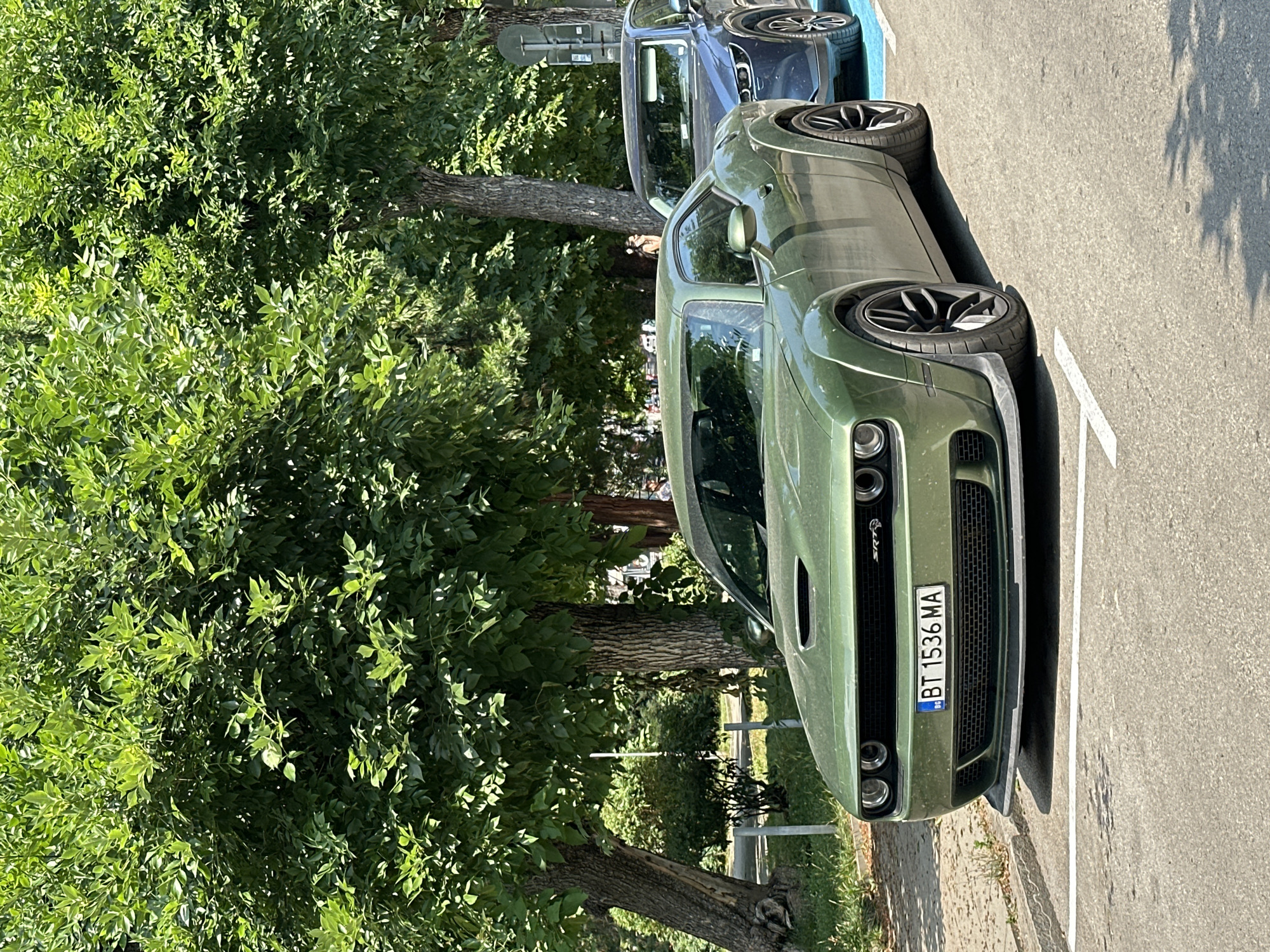 The image shows two parked cars in Veliko Tarnovo, Bulgaria. The primary subject is a green Dodge Challenger, facing right. It is parked on asphalt with white parking lines. The car has a prominent front grille and headlights. A license plate is visible on the front of the car, reading "BT 1536 MA" with a Bulgarian flag emblem. The car's side is visible, showcasing its wheels and body shape.
Behind the Dodge Challenger, a dark blue car is parked, also facing right. Only the rear of the blue car is significantly visible.
The setting appears to be outdoors, possibly a parking lot or street, with trees and foliage on the left side of the image. The trees are lush and green, indicating a warm season, likely daytime due to the bright sunlight casting shadows on the ground. The overall mood is calm and still, reflecting a typical daytime scene in a city. The presence of trees and greenery suggests a location that is not purely urban, perhaps a park or a tree-lined street. The time of day appears to be during daylight hours, with the sun casting distinct shadows from the trees.