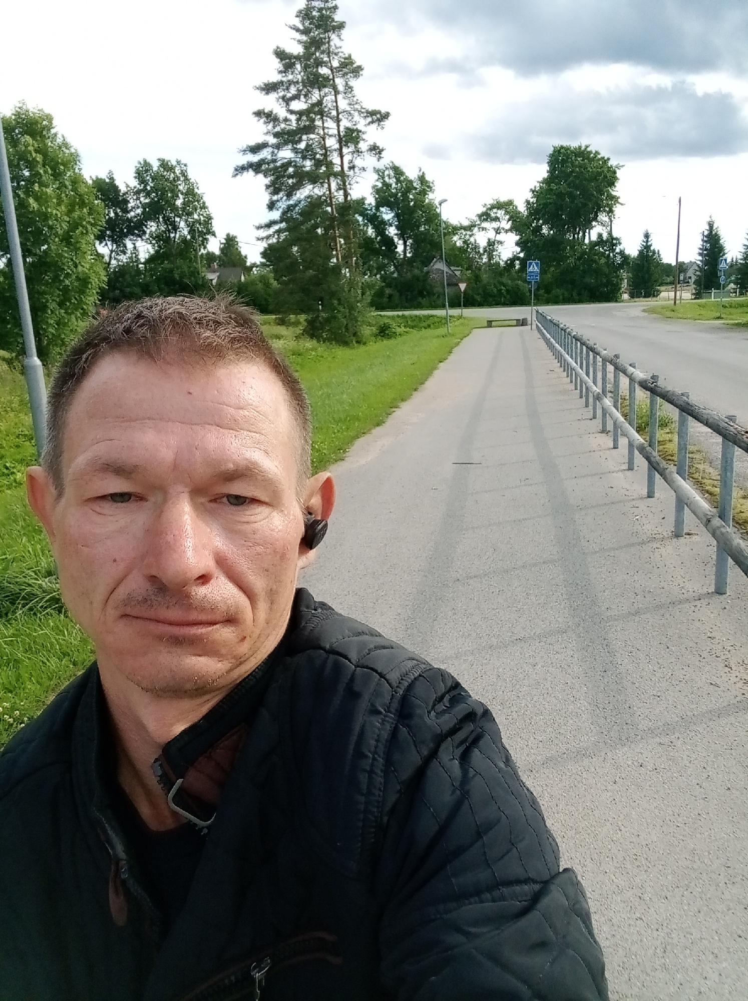 A man with short, graying hair and a light mustache stands on a paved walkway, looking directly at the camera. He is wearing a black, quilted jacket and a black earbud in his right ear. The walkway runs alongside a road bordered by a metal and wooden fence. Beyond the fence, there is a grassy area with trees and houses in the distance. The sky is overcast. The scene appears to be outdoors on a clear day. The photo was taken in Tapa, Estonia.