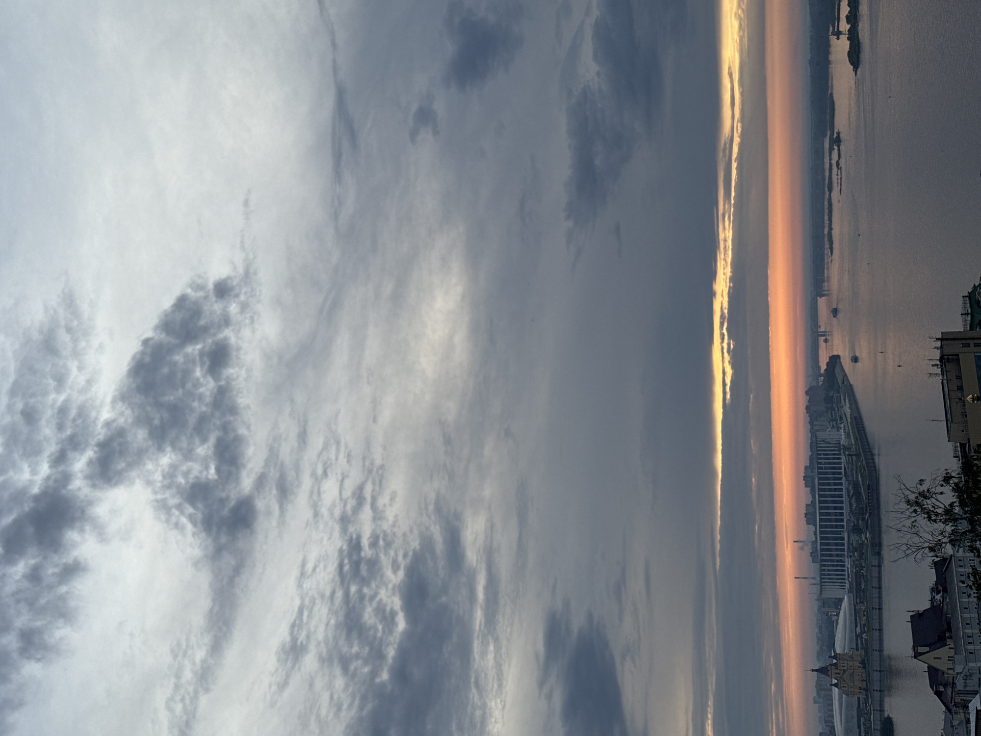 The image shows a sunset over Nizhny Novgorod, Russia. The sky is filled with gray and white clouds. A strip of orange light separates the clouds from the city and the Volga River. The river reflects the light of the sunset. Buildings and trees line the river bank.