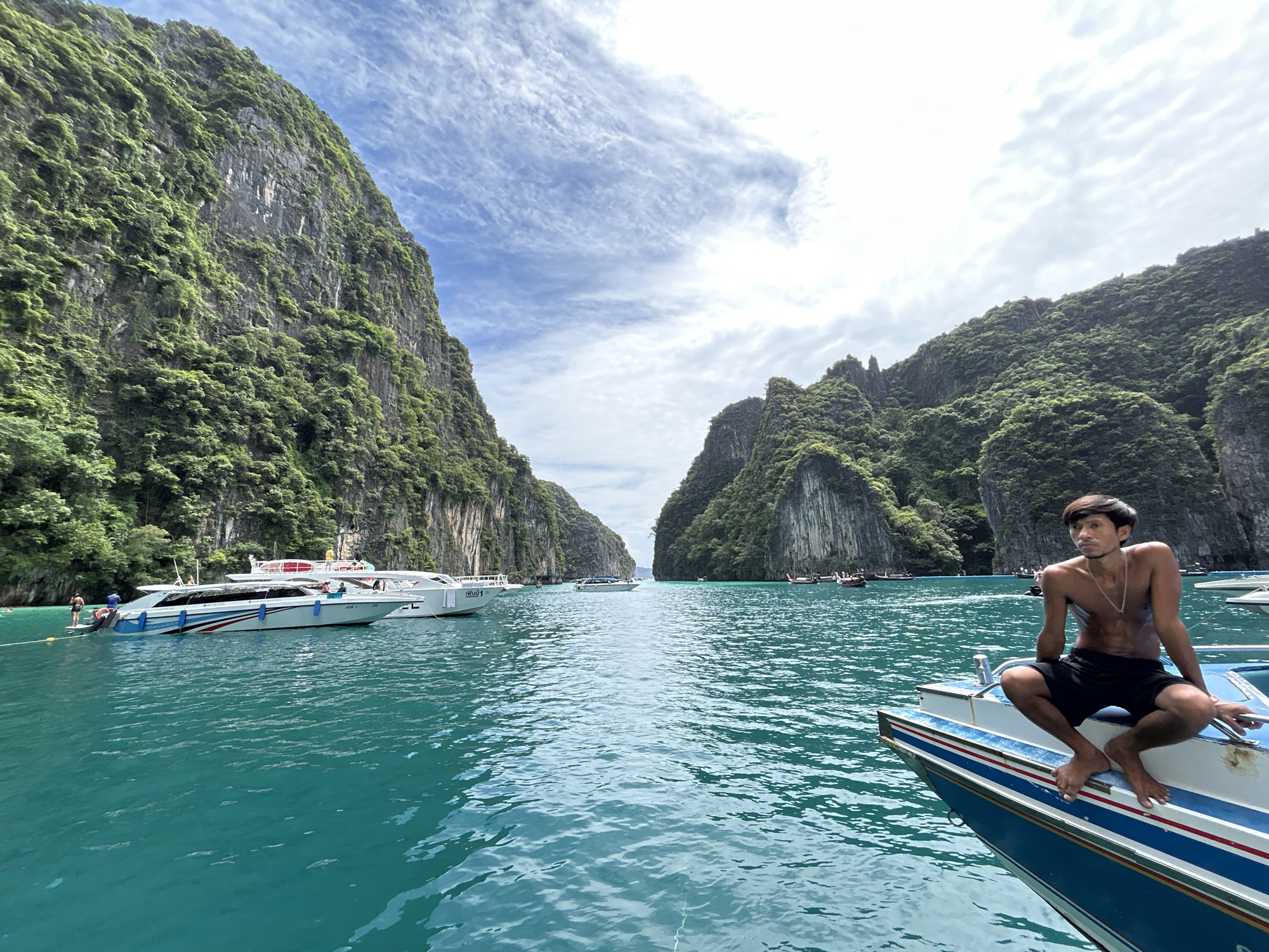 A young man with dark hair and a thin build, wearing black shorts and a silver necklace, is seated on the edge of a boat. He is looking to his left, with his right leg bent and his left leg extended. The boat is blue and white with red and green stripes. The water is a clear turquoise. In the background, there are towering, green, rocky cliffs that line a body of water. Several other boats, including speedboats and longtail boats, are visible in the distance. The sky is partly cloudy with streaks of white clouds against a bright blue sky. The overall scene depicts a tropical island setting in Thailand.