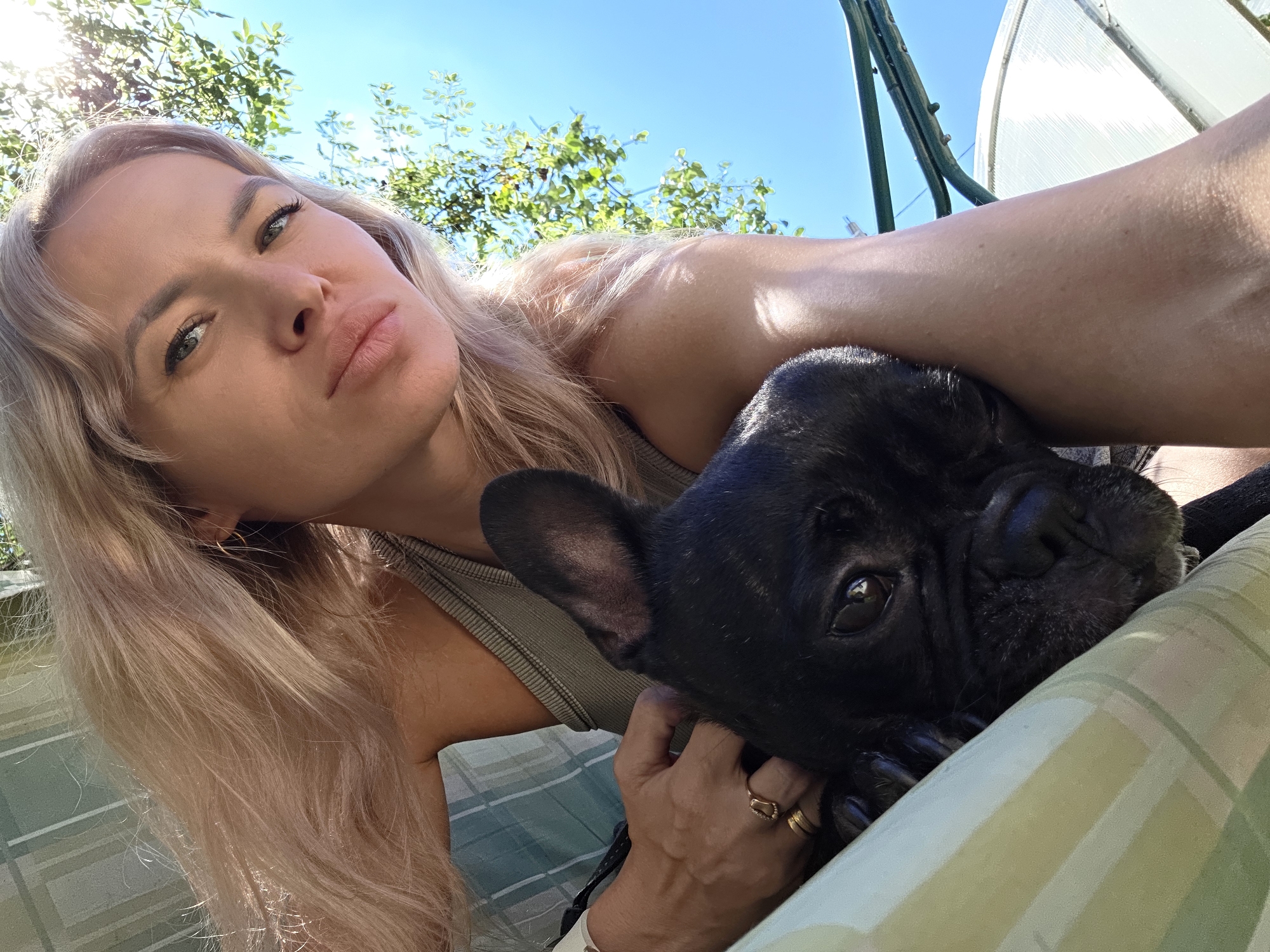 A woman with blonde hair and a black French bulldog are shown in a close-up, low-angle selfie. The woman is leaning towards the camera, with her left arm around the dog. She is wearing a grey tank top and gold rings. The dog is nestled against her, with its head resting on her arm. The background shows a bright blue sky, green foliage, and a white greenhouse. The scene appears to be outdoors in a backyard setting. The image was taken in Chaikovsky, Russia.