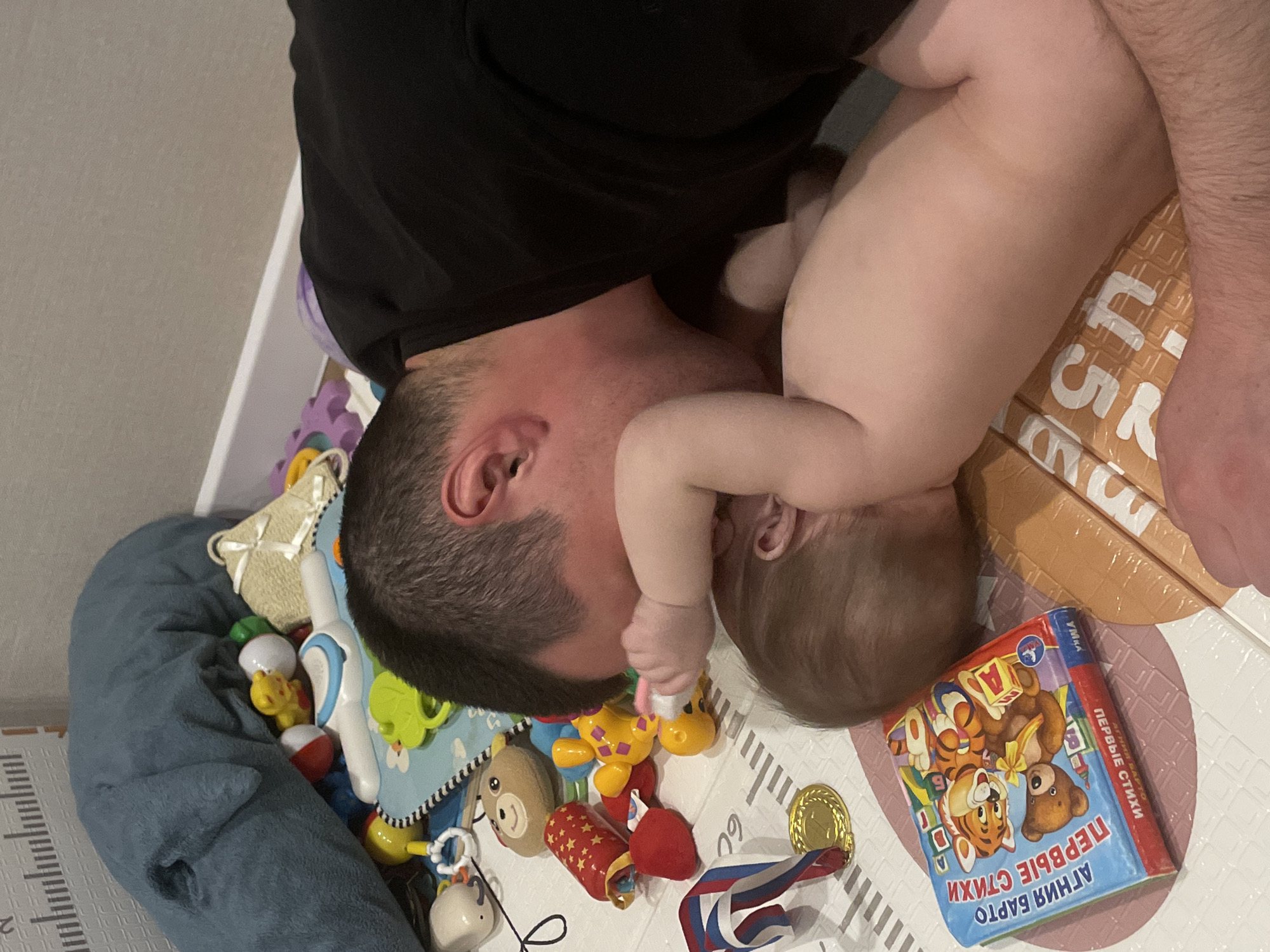 A man and a baby are lying on a play mat surrounded by toys. The man is wearing a black shirt. The baby is naked. A children's book titled "Первые стихи" is visible in the lower right corner. The scene appears to be indoors, possibly in a home in the city of Подшивалово, Russia, based on the book title.