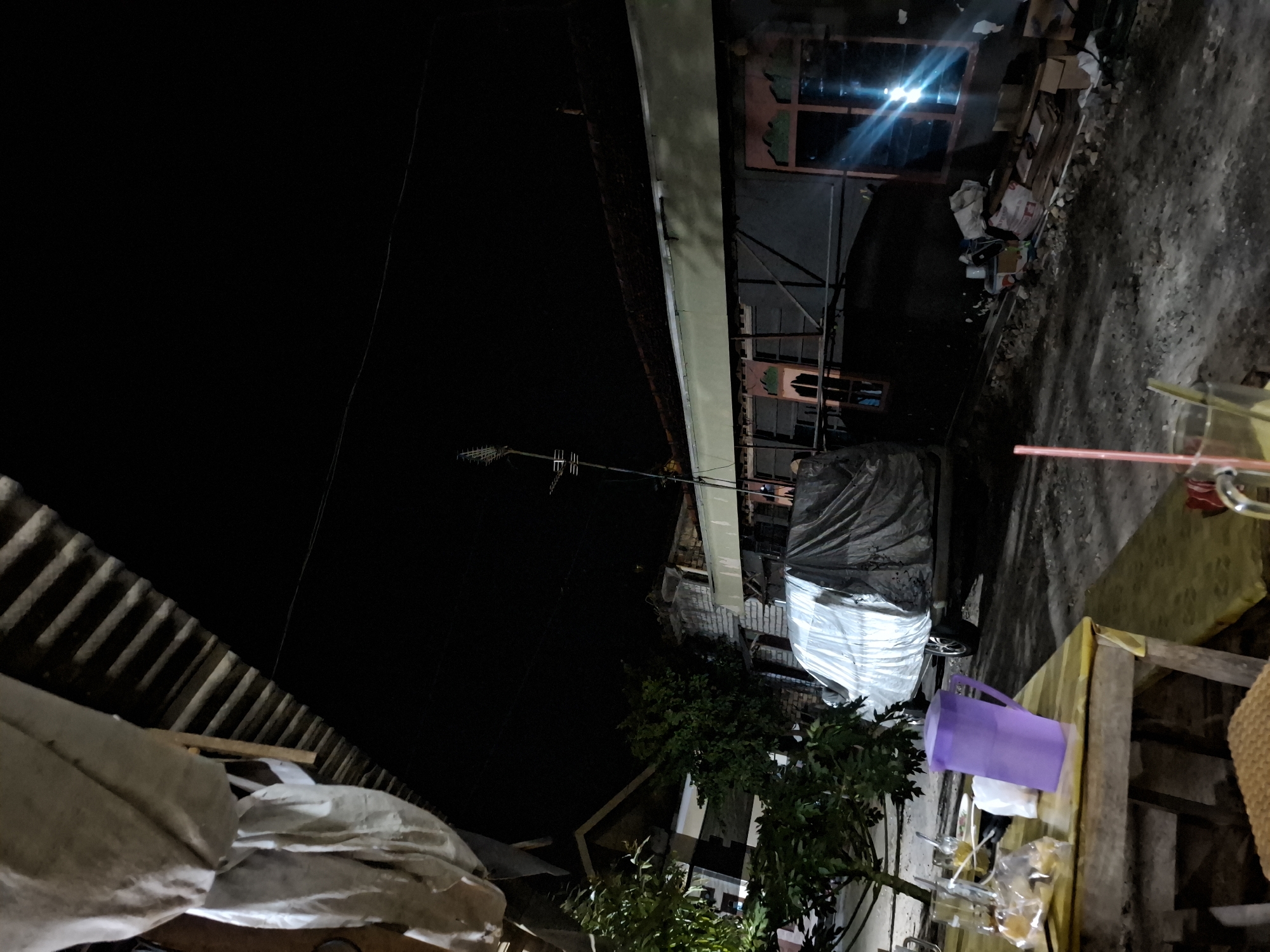In Bulubrangsi, Indonesia, this night-time scene captures a rustic outdoor setting. A wooden table in the foreground is laden with drinks in glasses, a purple pitcher, and some bagged items. Beyond the table, a silver van is partially covered by a tarp. The backdrop consists of a dark, overcast sky, the corrugated roof of a building, and a row of houses with visible windows, one of which reflects a bright light source. Trees, building structures, and debris are also present, contributing to the area's somewhat unkempt appearance.