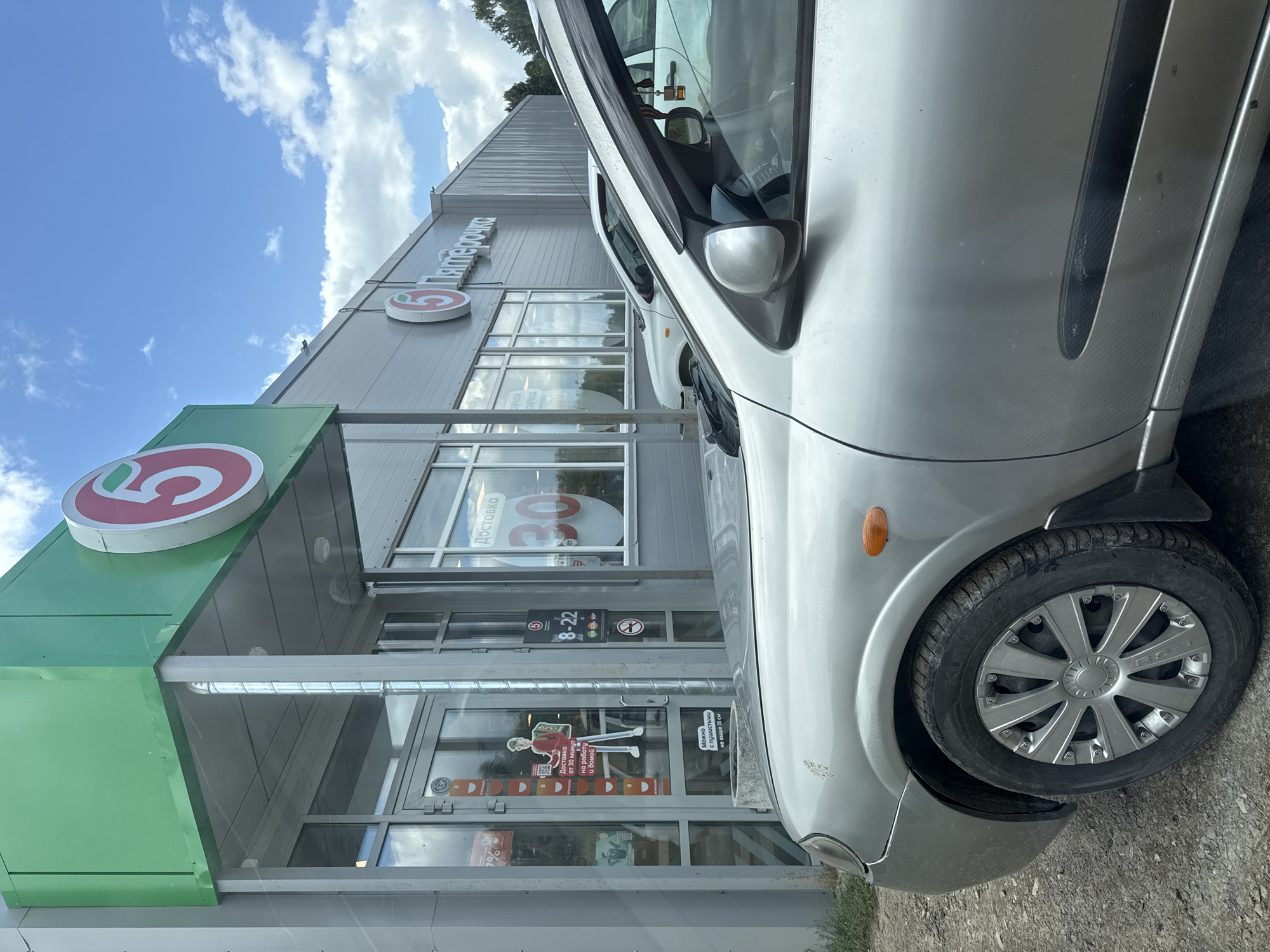 In an outdoor scene, a silver car is parked in front of a store with a green canopy and a large "5" logo. The store's signage reads "Пятёрочка" (Pyaterochka). The car is a close-up, showing its front fender, wheel with a silver hubcap, and part of the door. The store entrance features glass doors with several stickers and a sign indicating operating hours from 8 AM to 10 PM. A window behind the entrance displays a graphic with "Доставка 30" (Delivery 30) and "от" (from). Another car is parked in the background, and the sky above is blue with scattered white clouds. The ground is covered in gravel.