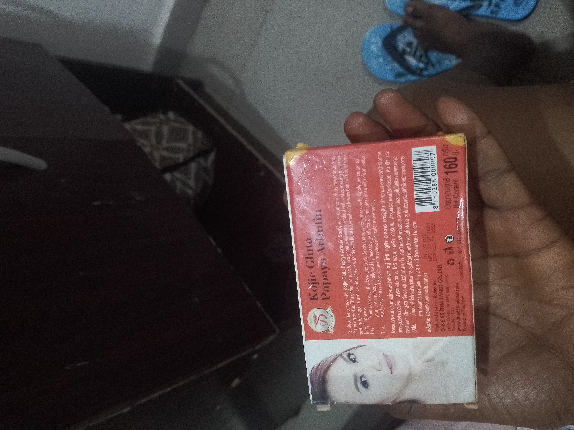The image is a close-up, overhead shot of a red box of Kojie Gluta Papaya Arbutin soap. The box is positioned at an angle on a dark surface, partially obscuring a patterned fabric and out-of-focus blue flip-flops in the background. The soap box has English text on one side detailing instructions for use, ingredients, and production information, along with Thai text and a barcode. A portion of a woman's face is visible on the bottom of the box. The lighting is dim, casting shadows and creating a slightly grainy effect. The image appears to be taken in an indoor setting.