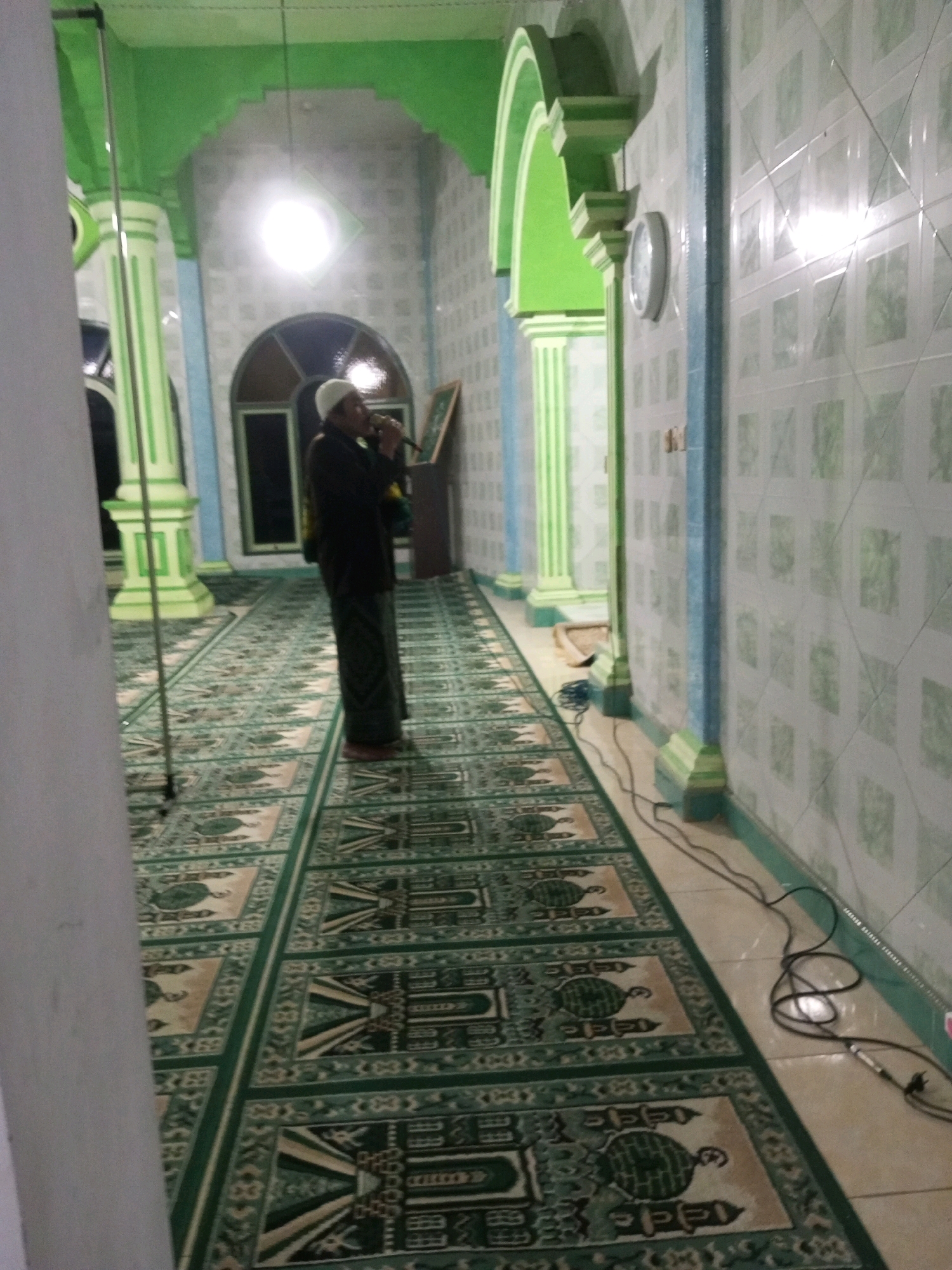 The image depicts the interior of a mosque with green and white tiled walls and green decorative arches. A man wearing a white skullcap and dark clothing, with a sarong around his waist, is standing in the middle of the prayer hall, holding a microphone to his mouth. He appears to be speaking or singing. The floor is covered with a carpet featuring a repeating pattern of green and beige geometric designs, possibly depicting prayer mats or mosque architecture. Several pillars, painted green and white, line the corridor. A clock is visible on the wall. Power cords are present on the floor near the wall. The lighting in the mosque is a mix of artificial light from a ceiling fixture and ambient light reflecting off the tiled surfaces. The overall impression is of a place of worship during a religious observance.