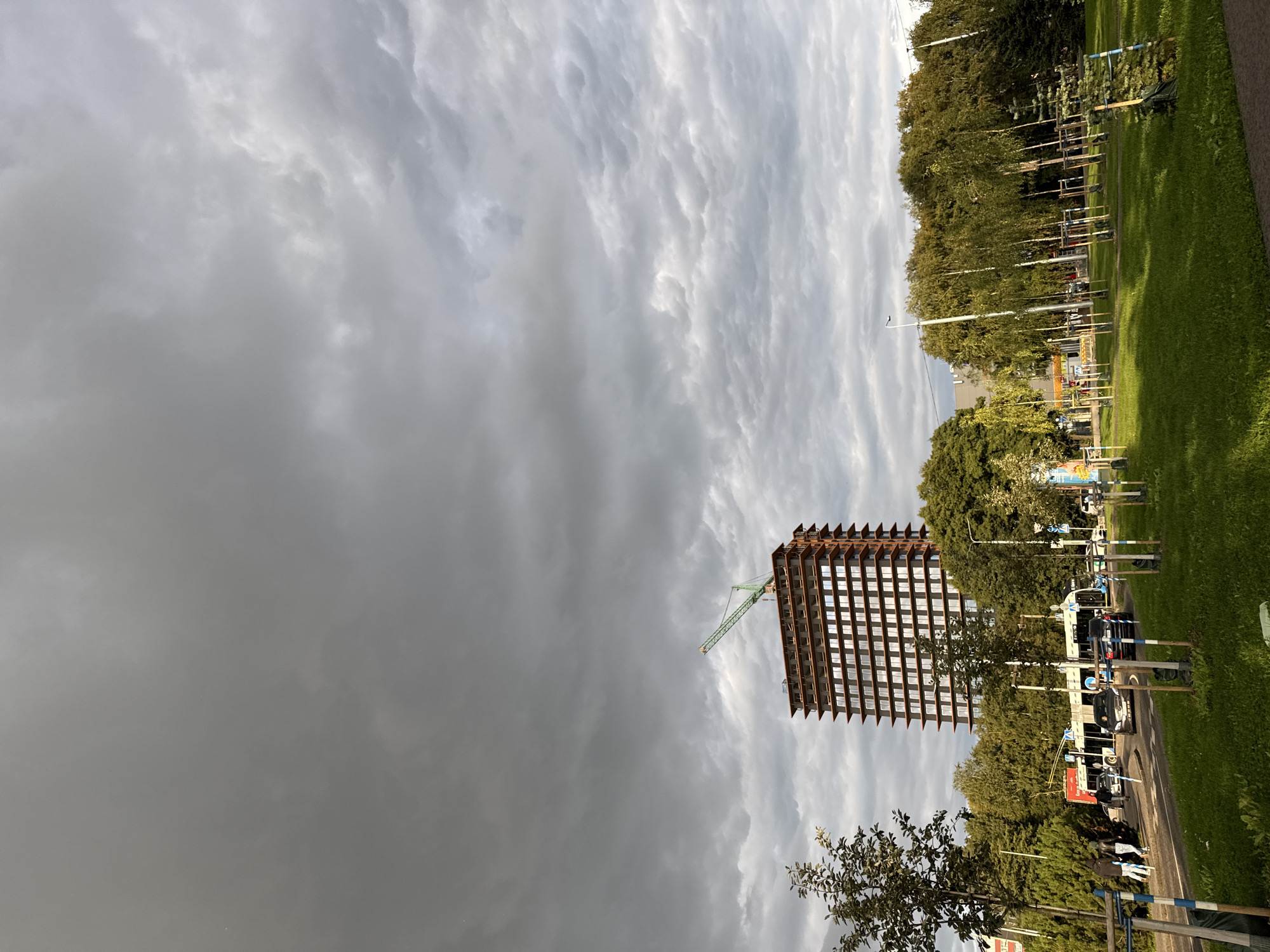 The image shows a construction site in Tallinn, Estonia, with a tall, unfinished building in the background topped with a green crane. The sky is overcast with dramatic, textured clouds. In the foreground, there is a grassy park with several young trees staked with wooden poles and blue and white markers. A street runs parallel to the park, with cars and a trolleybus visible. Several pedestrians are walking on the sidewalk near the street. The overall scene suggests a typical urban environment with ongoing development.