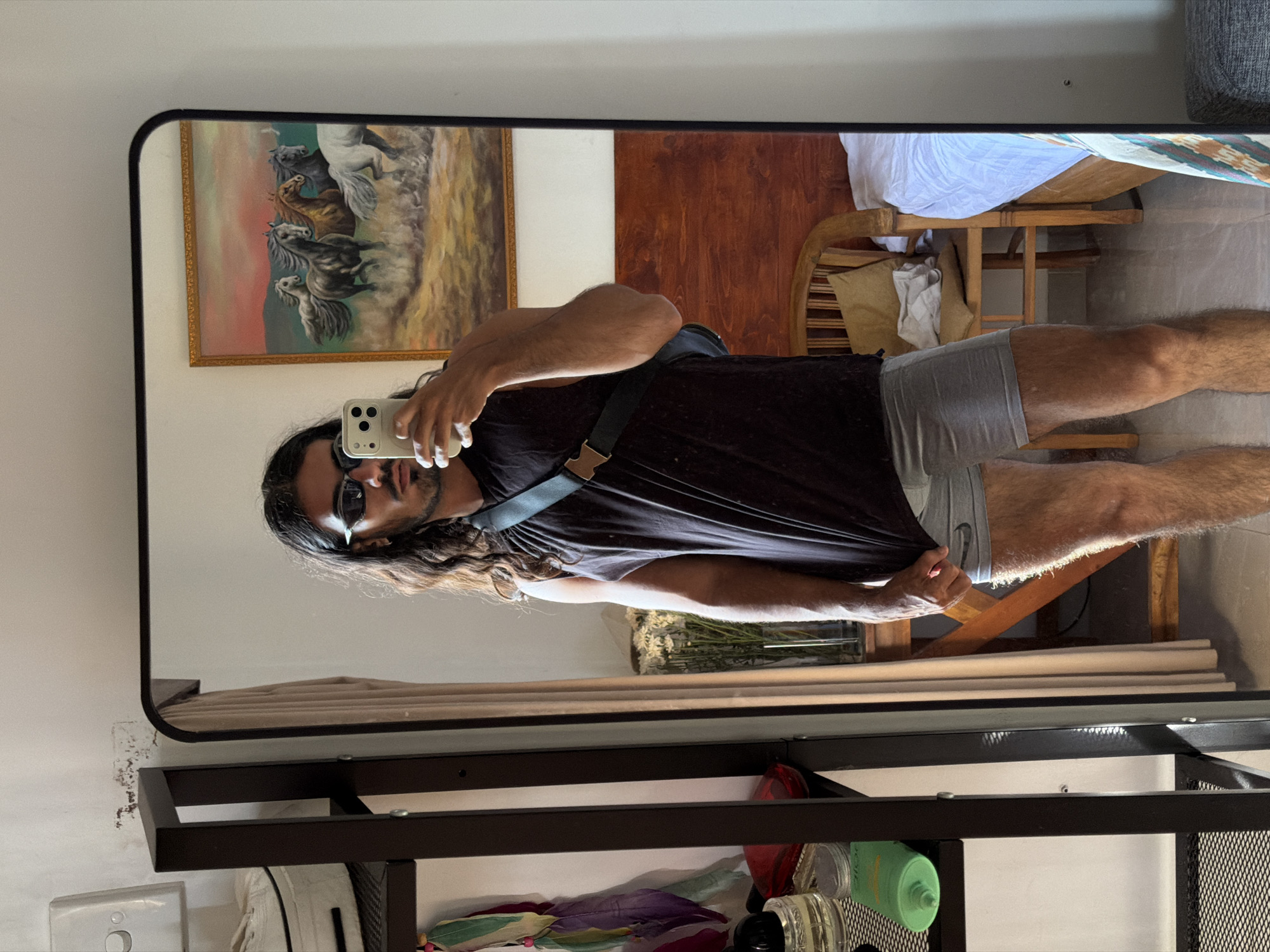 A person with long, dark hair, wearing sunglasses, a dark tank top, and gray shorts, is taking a selfie in a mirror. The person is holding a white smartphone. The reflection in the mirror shows a painting of horses, a wooden headboard and chair, and a partially visible bed with white linens. The mirror is mounted on a black metal shelf unit that contains various items. The location is North Kuta, Indonesia.