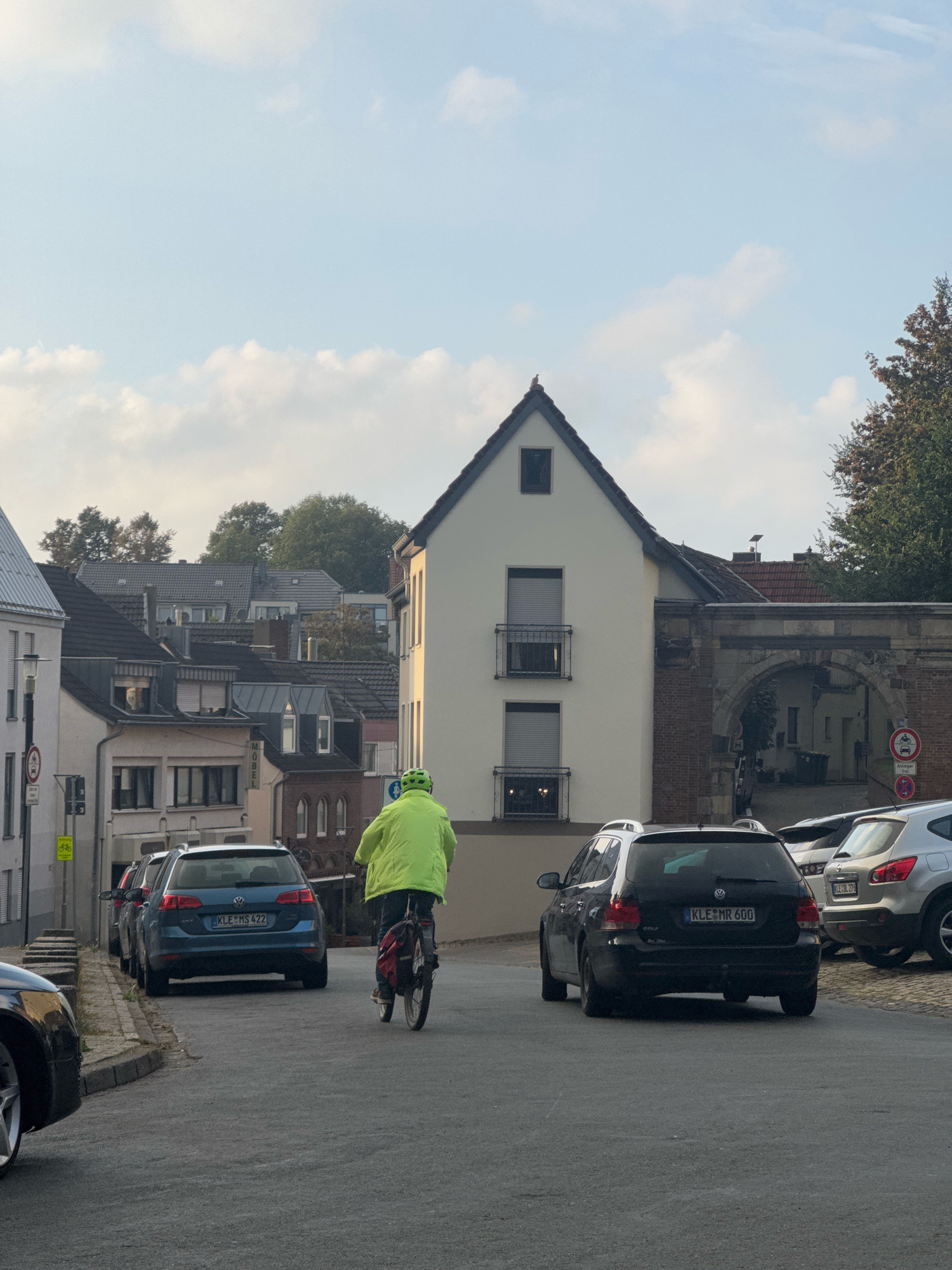 In the city of Cleves, Germany, a man on a bicycle wearing a bright yellow jacket and helmet is riding down a street. The bicycle has a red backpack on the rear rack. The street is paved and slopes downwards. Several cars are parked along the side of the street and on the right side of the road. The buildings lining the street are typical of a European town, with pitched roofs and varying architectural styles. A prominent white building with a dark roof and two balconies is visible in the center of the image. An arched stone structure is attached to this building. The sky is partially cloudy. Traffic signs are visible on the left side of the street. The license plate on a blue Volkswagen Golf is visible and reads "KLE MS 422". The license plate on a black Volkswagen Golf reads "KLE MR 600".