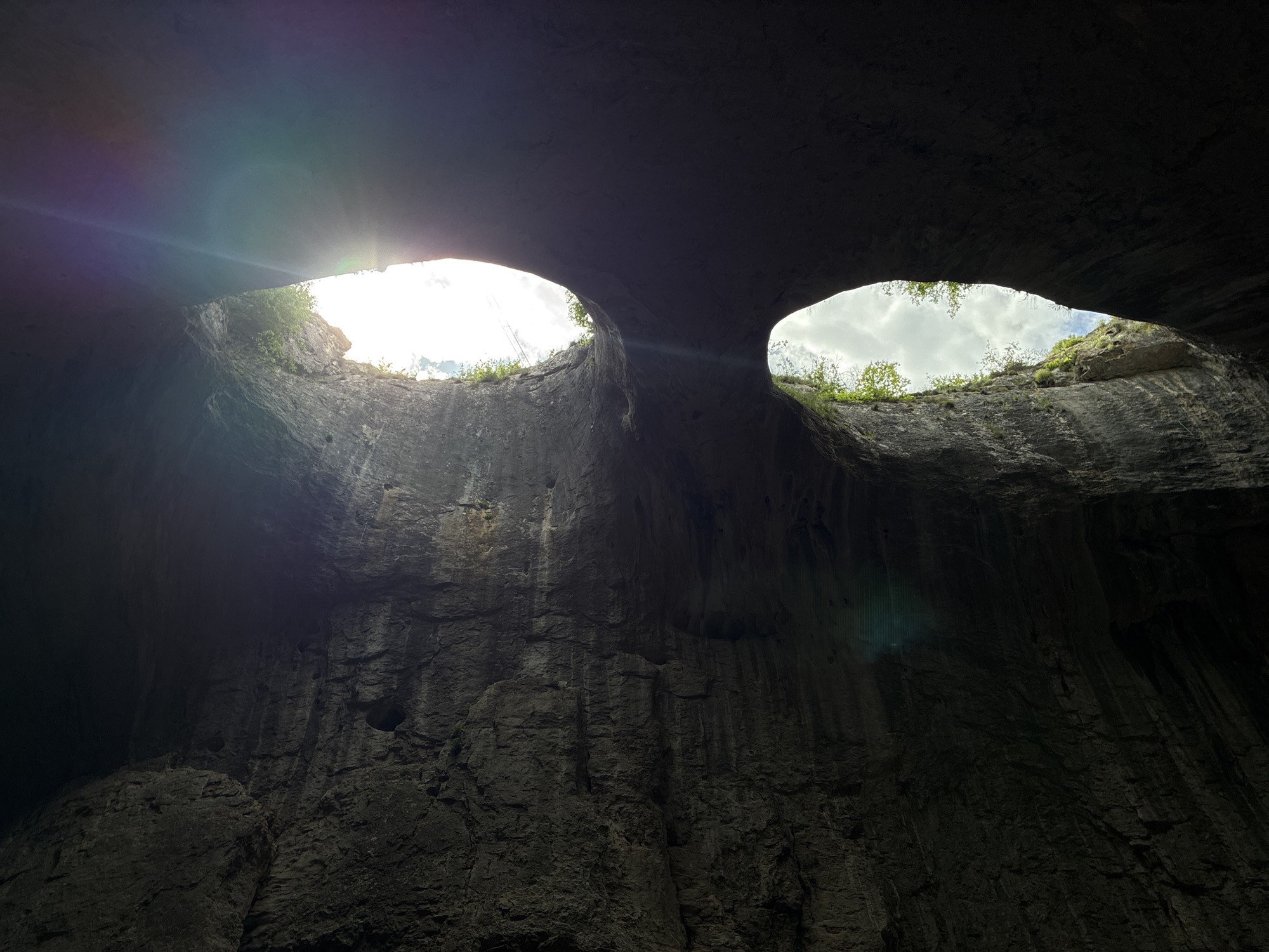 The image shows the interior of Devetashka Cave near Karlukovo, Bulgaria. Two large openings in the cave roof allow daylight to enter. The opening on the left is brighter, showing intense sunlight, while the opening on the right reveals cloudy skies and some vegetation. The cave walls are dark, textured with rock formations and vertical striations. The scene emphasizes the contrast between the darkness inside the cave and the bright light entering through the openings.