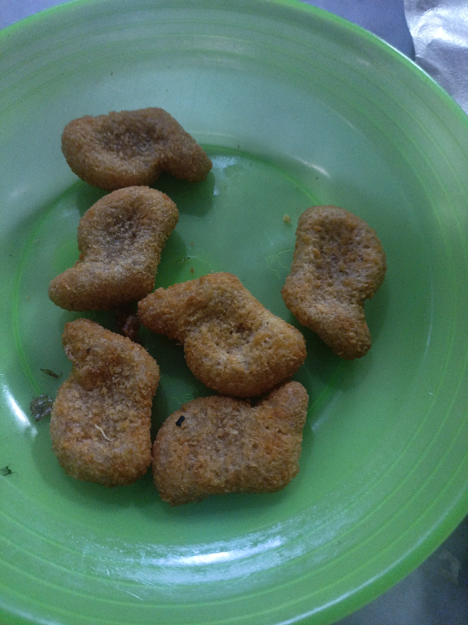 This is a close-up, overhead shot of several fried chicken nuggets arranged on a bright green plastic plate. The nuggets are irregular in shape, with a crispy, golden-brown breading. Some of the nuggets have a slightly elongated or curved form. The plate has visible concentric rings and appears to be slightly worn. The background is mostly out of focus, with hints of a gray surface. The lighting suggests an indoor setting. There are no people visible in the image. The scene is a food presentation, likely for a meal. It is not possible to determine the location in Undaan, Indonesia from the image alone.
