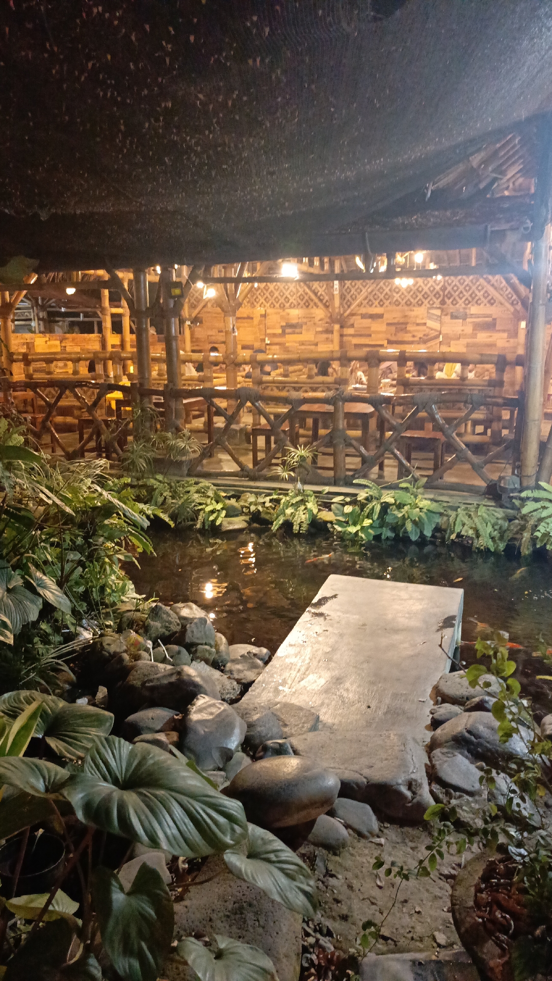 This is a medium shot, vertically oriented, night-time photograph of an outdoor seating area adjacent to a pond with a bamboo-themed aesthetic. The foreground features a shallow, dark pond with visible, small orange fish swimming near the surface. A concrete slab acts as a small bridge or walkway over the pond. Surrounding the pond is lush green foliage, including large-leafed plants and ferns, interspersed with large, smooth grey rocks.
The background reveals a covered seating area, constructed primarily from bamboo poles and woven bamboo panels, creating an intricate wall pattern. Numerous small, warm-toned lights are suspended from the bamboo ceiling, illuminating the dining tables and chairs arranged within the covered space. The overall impression is of a tranquil, rustic, and possibly dining establishment in Pringsewu, Indonesia. No people are clearly identifiable in the scene.
