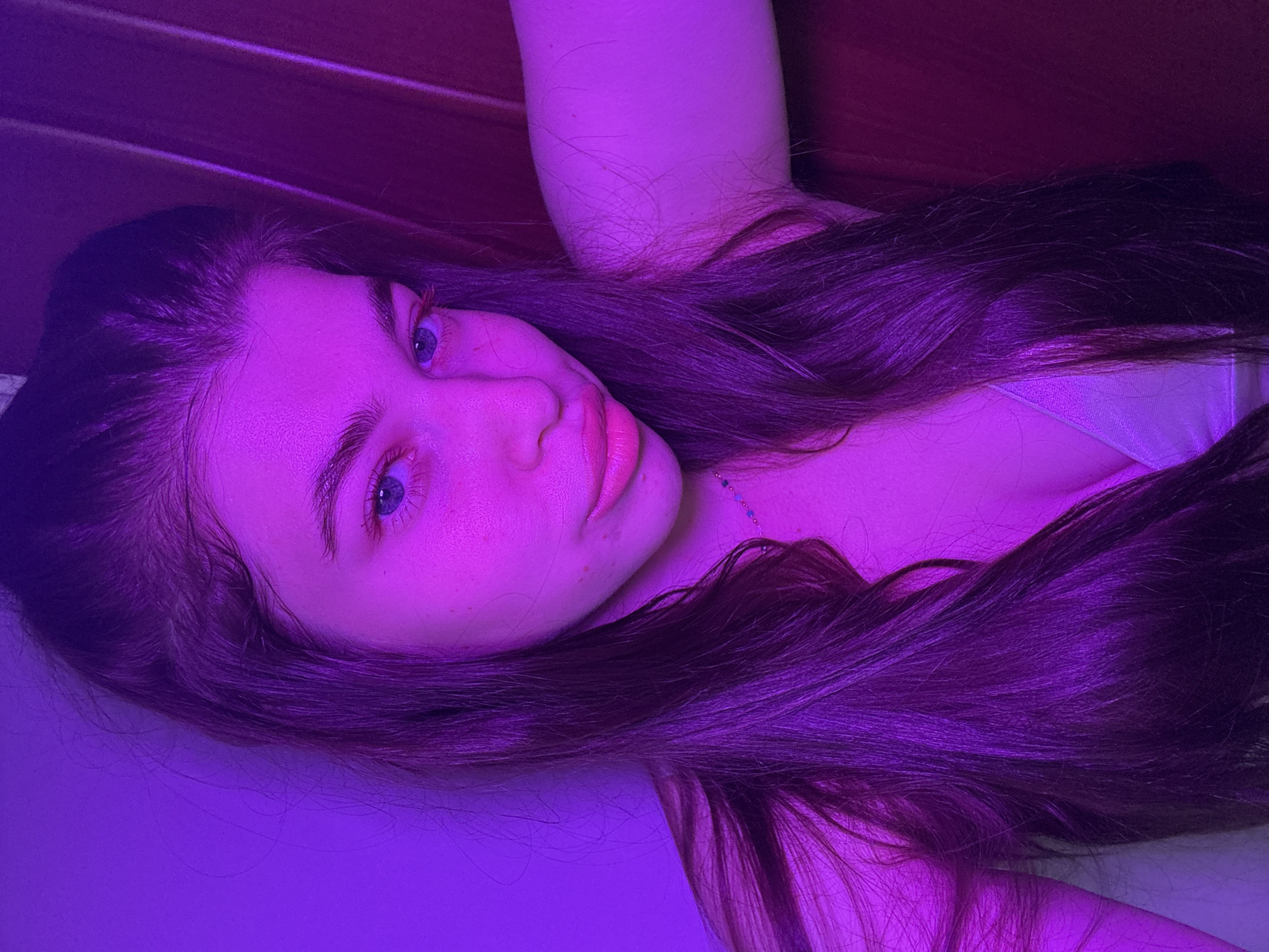 A young woman is pictured from a close, angled perspective. She is lying down with her head near a wooden-paneled surface. Her long, dark hair is spread around her. Her skin is pale. She has light eyes and is looking directly at the camera. The entire image is lit with a strong purple hue, casting an unnatural color across all elements of the scene. There is no indication of this image being taken in Мещёры, Russia.