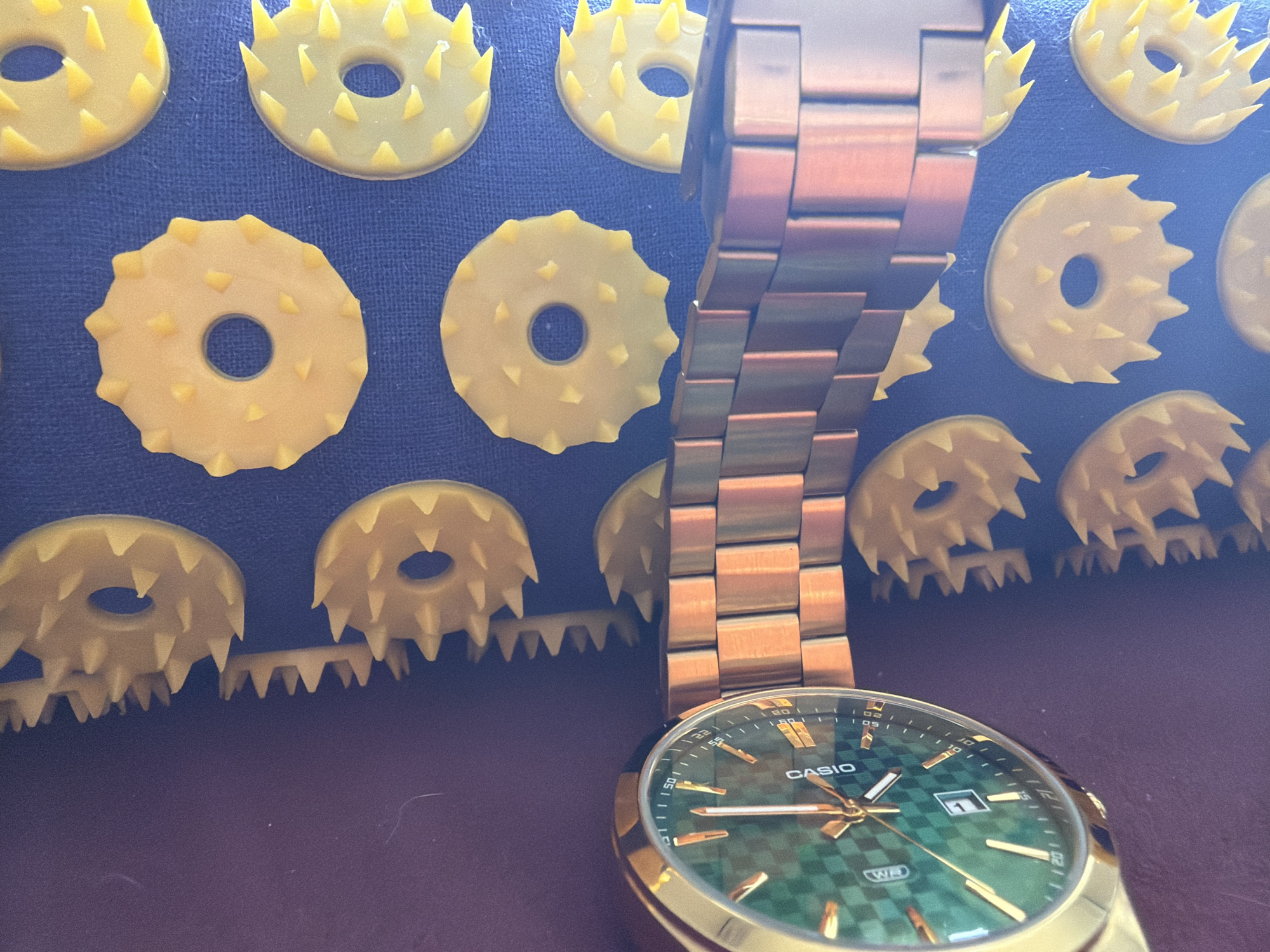 The image presents a close-up of a gold-toned Casio wristwatch with a metallic link bracelet, positioned on a dark, reflective surface. The watch features a striking green face with a checkered pattern, complemented by golden hour markers and hands. A date window displaying "1" is visible, along with text on the dial reading "CASIO" and "WR" (Water Resistant), surrounded by minute/second indicators.
Behind and partially beneath the watch, a textured background is formed by numerous yellow, spiky, circular objects. These objects resemble gears or components of an acupressure mat, characterized by sharp, conical protrusions along their edges and a central hole. They are arranged in a repeating pattern on a dark blue-purplish fabric or mat, with a horizontal strip of smaller, connected spiky elements visible in the lower foreground.
The scene is brightly lit, suggesting an indoor setting during the daytime, with reflections visible on the watch's metallic surfaces and the underlying dark platform. The overall composition highlights the watch as the primary subject against the distinct patterned background. This display is set in Chaikovsky, Russia.