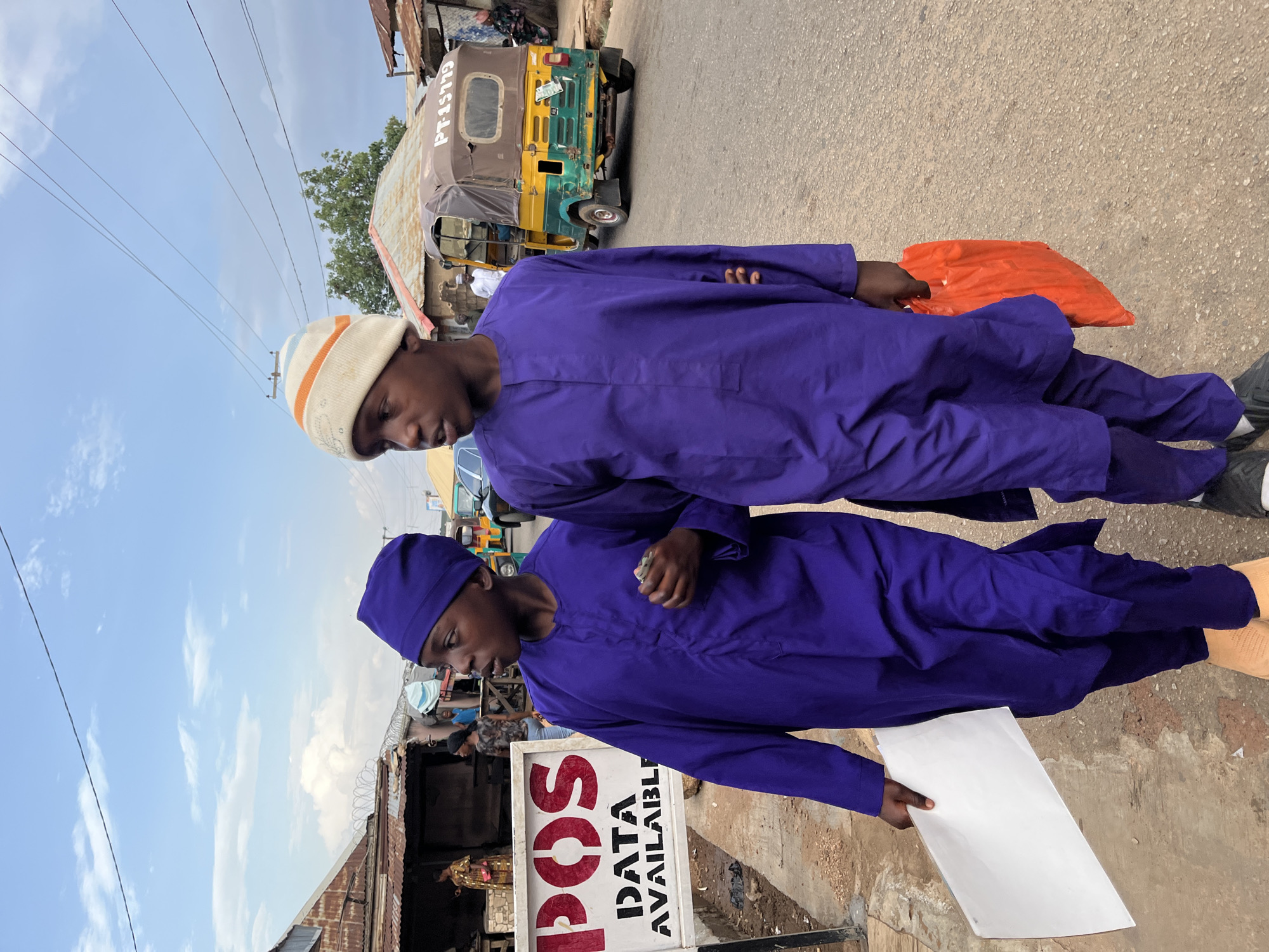 Two young men wearing matching purple attire stand next to a "POS Data Available" sign in Jos, Nigeria. The man closer to the sign holds a piece of white paper, while the other carries an orange bag. A vehicle, possibly a tricycle taxi, is parked on the street in the background. The setting appears to be an urban street with overhead power lines and nearby buildings.