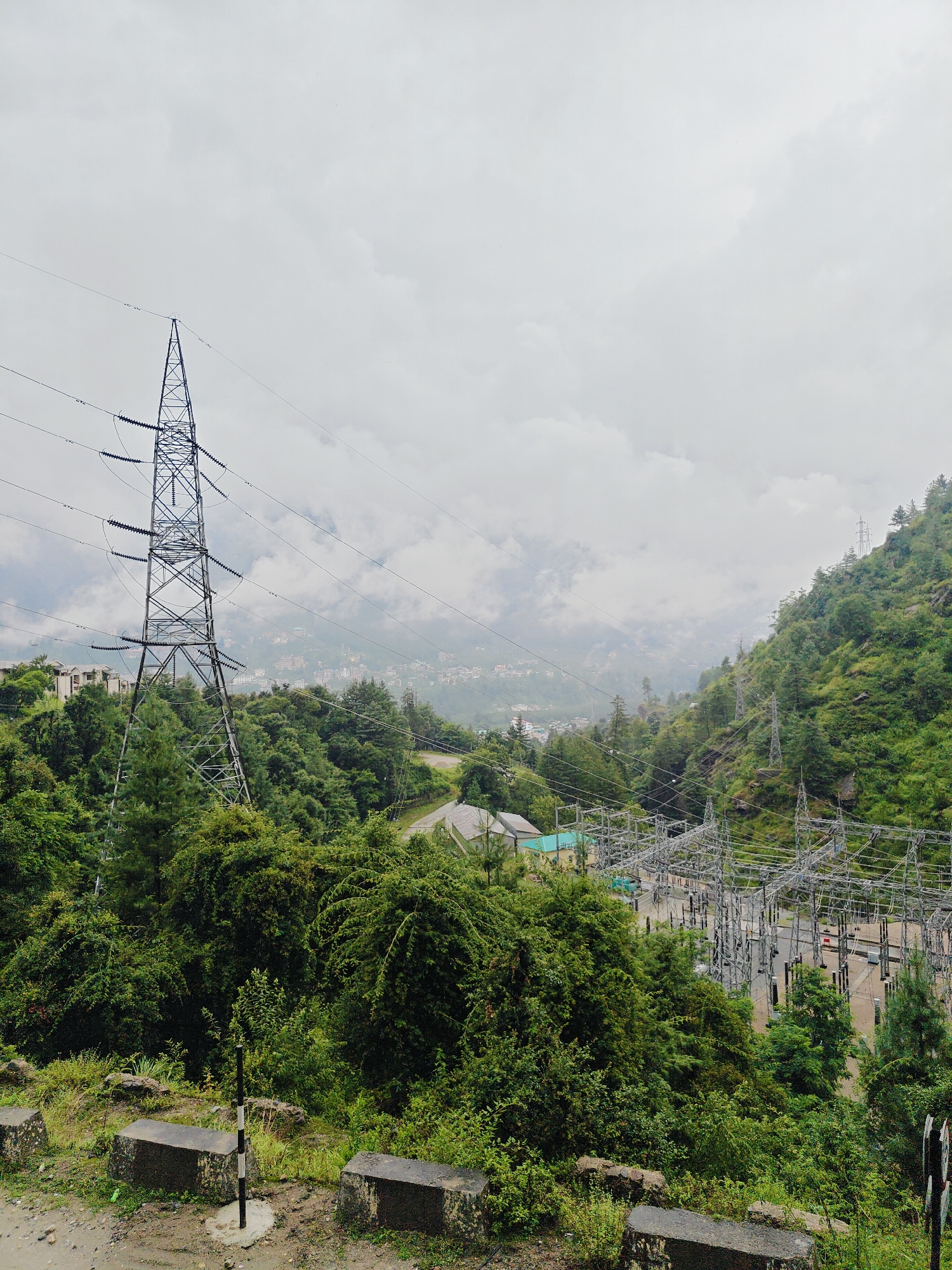 The image shows a power substation in Prini, India, nestled within a lush green, mountainous landscape. In the foreground, to the left, a tall, lattice-steel electricity pylon stands prominently, with high-voltage wires extending from it. Below the pylon, dense green vegetation covers the hillside. To the right of the pylon, a wide expanse of electrical infrastructure, consisting of numerous pylons, transformers, and insulators, is visible, spread across a cleared area. In the background, a town with buildings is spread across the slopes of the mountains, partially obscured by mist and clouds. The sky is overcast with grey clouds. The ground in the immediate foreground appears to be a dirt or gravel path with some concrete barriers.