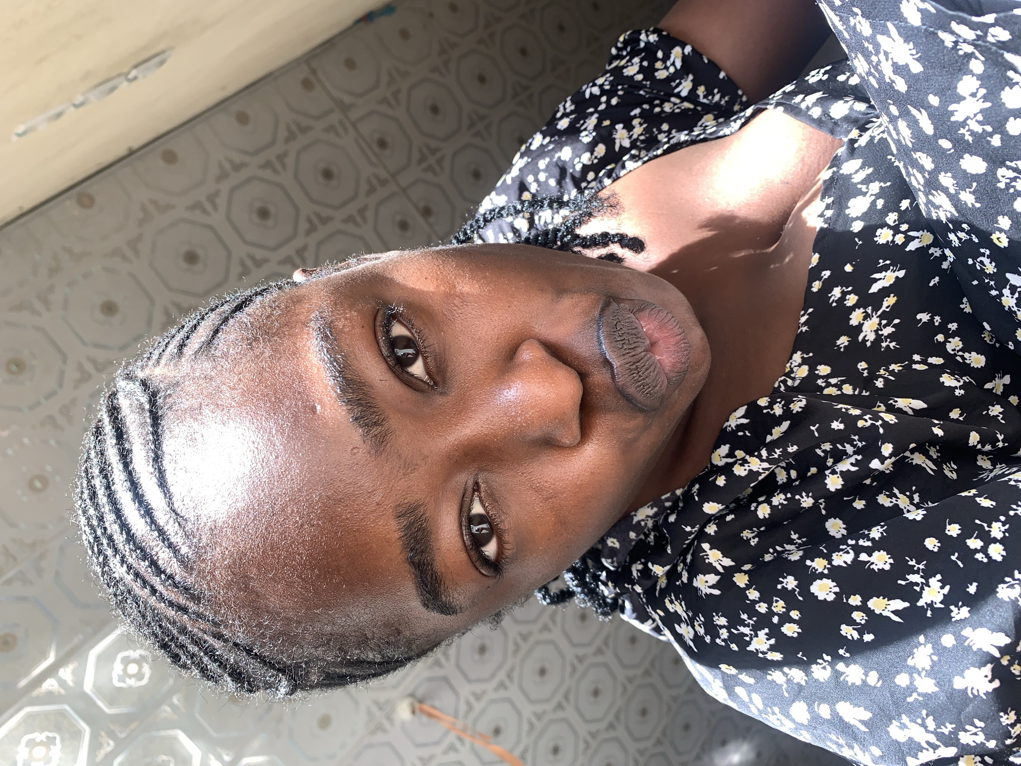 A close-up portrait features a person with dark skin and cornrow braids, looking directly at the camera with a neutral expression and slightly pursed lips. The person's head is positioned towards the left of the frame, with their body extending towards the right. They are wearing a black top adorned with small white and yellow floral patterns.
Strong, natural sunlight illuminates their face and clothing, creating bright highlights and distinct shadows. This suggests a sunny daytime setting, and the person's skin appears somewhat dewy, possibly from warmth. The background consists of light-colored, patterned floor tiles visible to the left and upper right of the person, indicating an indoor location. A plain, light-colored surface, possibly a wall or ceiling, is visible in the upper part of the frame. There is no visible text in the image. The location context for this image is New Stadium, Nigeria.