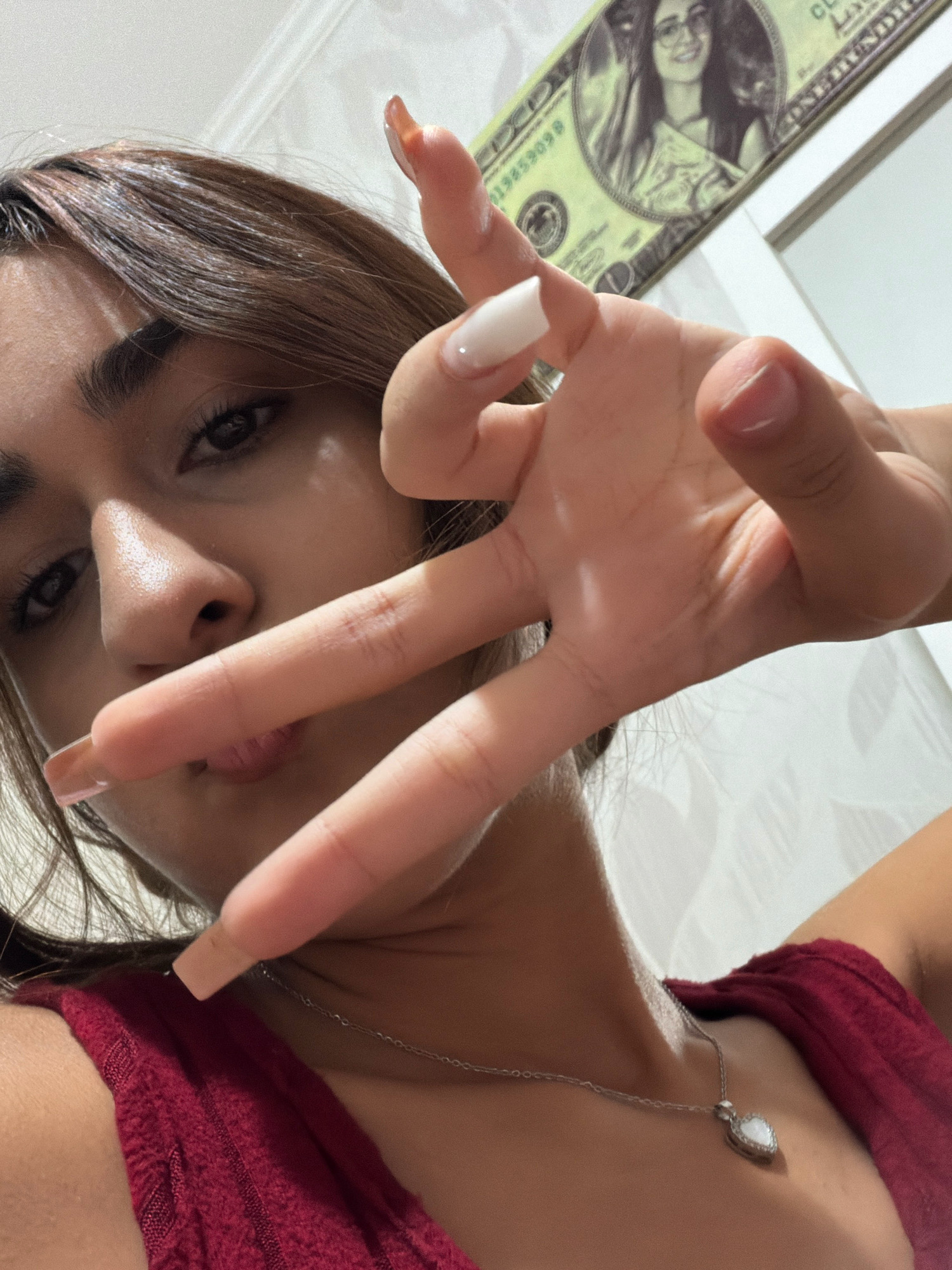 A young woman with brown hair and a red top is taking a selfie. She is holding up her hand in a peace sign, with her fingers in focus. In the background, a stack of bills with a woman's face on them can be seen. The woman is wearing a silver necklace with a heart-shaped pendant. The image appears to have been taken indoors, possibly in a bedroom, with white walls and some sort of patterned wallpaper visible. There is no information in the image to suggest the location is in Digah, Azerbaijan.
