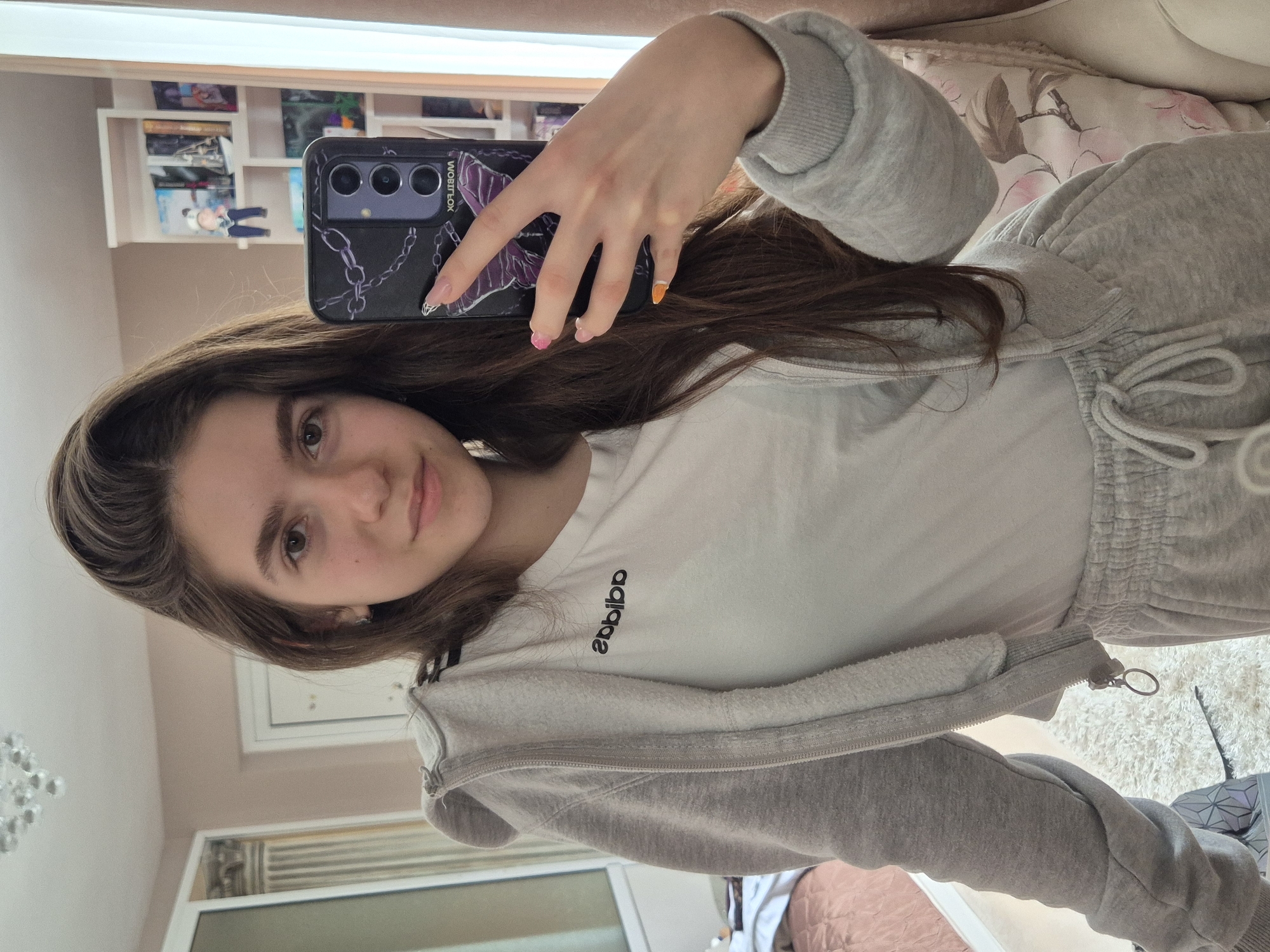 A young woman is taking a selfie indoors, holding a black smartphone with a decorative case. She is wearing a white T-shirt with the Adidas logo and a gray zip-up hoodie and sweatpants. Her nails are painted with different colors. Behind her are shelves with various objects, including books and figurines. A chandelier is visible in the background. The location is not identifiable as being in Pavlikeni, Bulgaria.